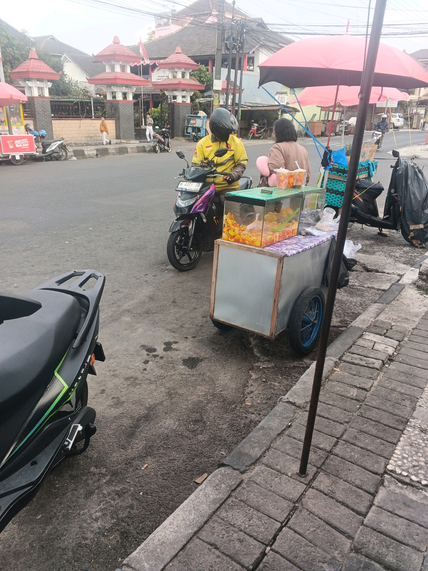 This image captures a street scene in Ungaran, Indonesia, near a building with distinctive red-roofed architectural elements and a fenced perimeter. In the foreground, a person wearing a yellow jacket and black helmet is seated on a purple and black scooter. Next to the scooter is a food cart with a glass display showcasing various fruits and snacks. A woman in a brown top stands behind the food cart, interacting with the scene. Two large, pink umbrellas are set up over the cart. Further down the street, several other scooters and a motorcycle are parked or in motion, and a building with a sign is visible in the background. The pavement is a mix of asphalt and brick. The overall atmosphere suggests a bustling local market or roadside vendor area.