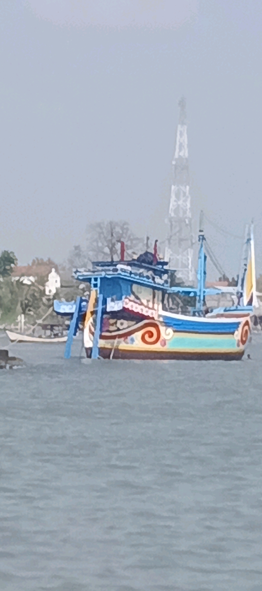 In the city of Blimbing, Indonesia, a brightly colored fishing boat with intricate swirling patterns adorns the water, its blue and yellow hues standing out against the muted tones of the overcast sky and the grey waves. The boat is equipped with multiple masts and rigging. In the background, a distant, lattice-like tower pierces the hazy atmosphere, and a cluster of buildings, some with white walls and red roofs, dot the shoreline, partially obscured by trees. Another small, white boat is visible closer to the shore. The scene conveys a sense of a coastal community engaged in its daily activities, with the fishing boat a prominent symbol of its maritime heritage.