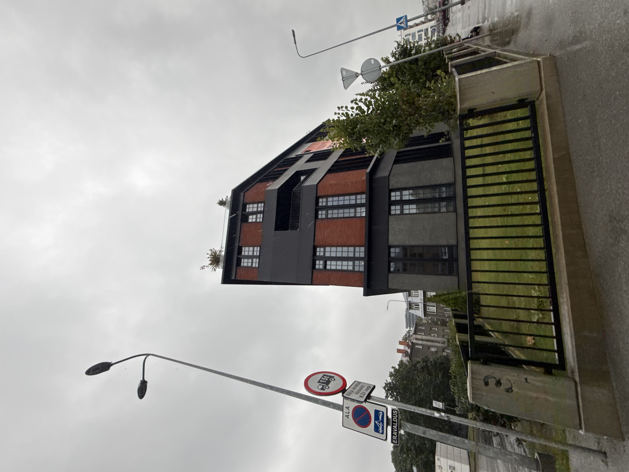 The image captures a street view in Tallinn, Estonia on a cloudy day, with a modern brick and dark-paneled building dominating the frame. The building features multiple stories with large, dark-framed windows and a balcony on one of the upper levels. A small tree is visible on the roof of the building. In the foreground, a metal fence encloses a patch of grass. To the left, a street light with two lamps stands tall, next to a cluster of traffic signs. One sign indicates "ALA" with a no parking symbol and a car icon, another sign shows a truck with "15 t" circled in red, and a rectangular sign reads "ERAVALDUS," meaning "Private property." Further down the street, other buildings and trees are visible. The road surface appears wet, suggesting recent rain. The overall scene is of an urban environment with a mix of modern and older architecture.