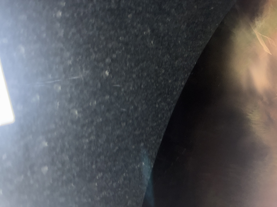 The scene is located in La Marquesa, Mexico. The image presents an obscured view, horizontally bisected by a curved line. The upper two-thirds of the frame consist of a dark, textured surface, possibly charcoal gray or black, uniformly covered with numerous small, irregularly shaped, light-colored particles, suggesting dust or debris. A bright, rectangular light source, cropped, is visible at the very top edge. Faint, thin, vertical linear marks are discernible on this upper surface. The lower third of the frame reveals a smooth, dark, curved surface, which reflects a heavily blurred and out-of-focus external scene. This reflection is characterized by diffuse warm tones, including shades of orange, yellow, and reddish hues, along with vague, elongated, lighter patterns. No identifiable people, distinct objects, or specific actions are visible within the frame.
