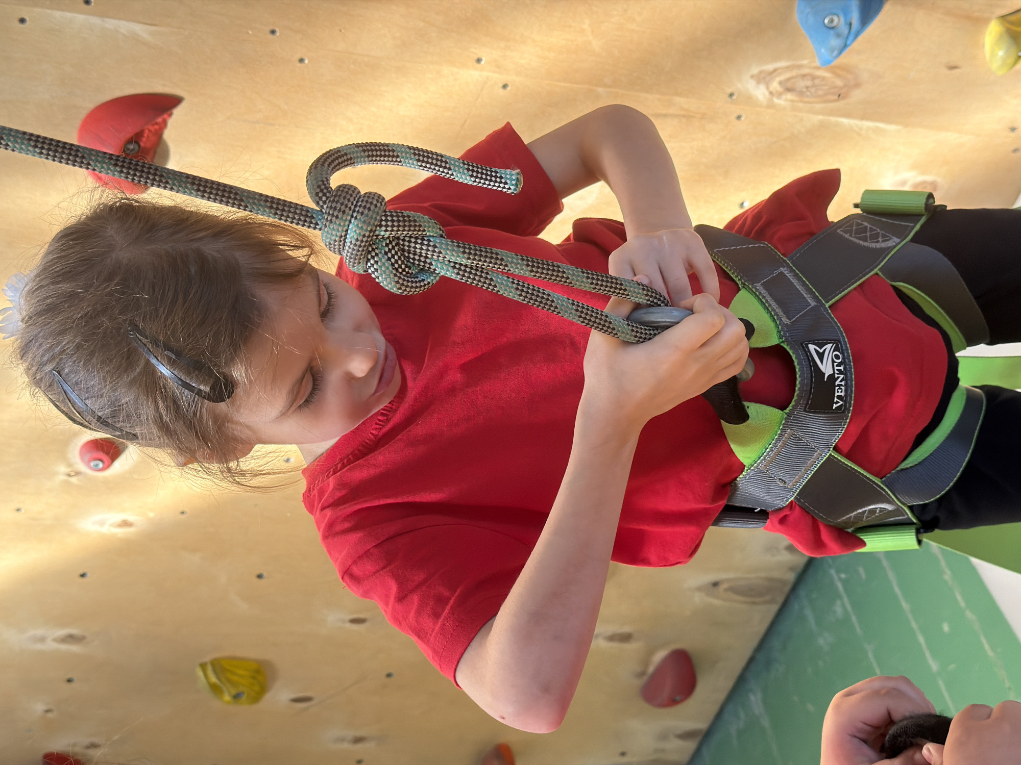 A young person wearing a red t-shirt and a climbing harness is holding a rope on a climbing wall. The wall is made of light-colored wood and has climbing holds attached to it. The rope is tied in a knot. The brand "VENZO" is visible on the harness. The location and the city of Izhevsk, Russia, cannot be verified from the image.