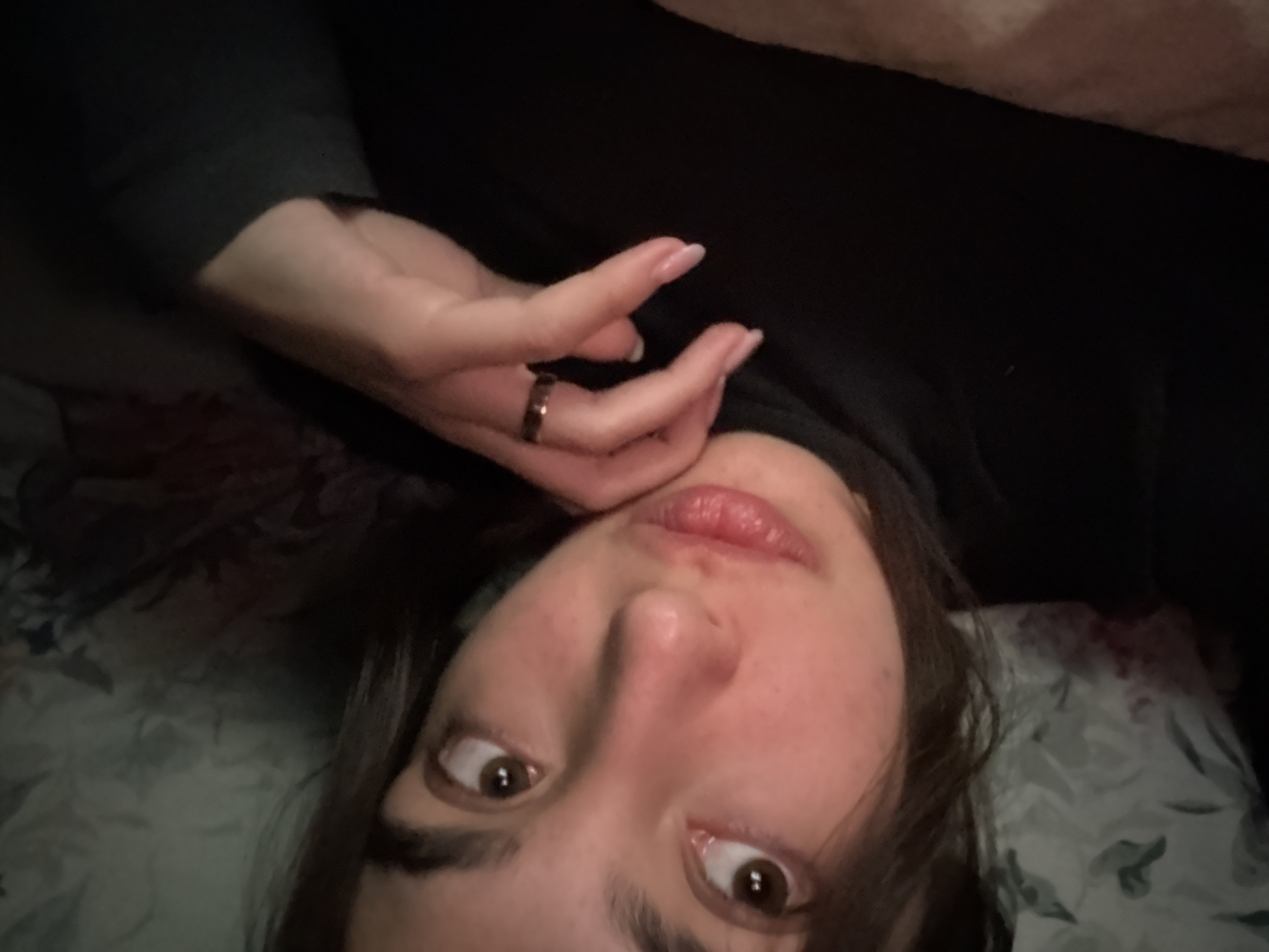 A close-up, low-light portrait captures a person, likely a young woman, lying down and looking directly into the camera. Her face is presented upside down from the viewer's perspective, framed by dark hair. She has wide, dark brown eyes and slightly parted lips. One hand is positioned near her chin, with a dark-colored ring visible on her right ring finger. Her fingernails are manicured with light tips. She appears to be wearing a dark, long-sleeved garment. The background is dimly lit, showing patterned fabric, possibly bedding with a subtle botanical design, on the left side of the frame, and plain dark fabric on the right. The scene suggests an indoor, intimate setting, possibly a bedroom, during the evening or night. Her expression conveys curiosity or slight surprise. No text is visible in the image.