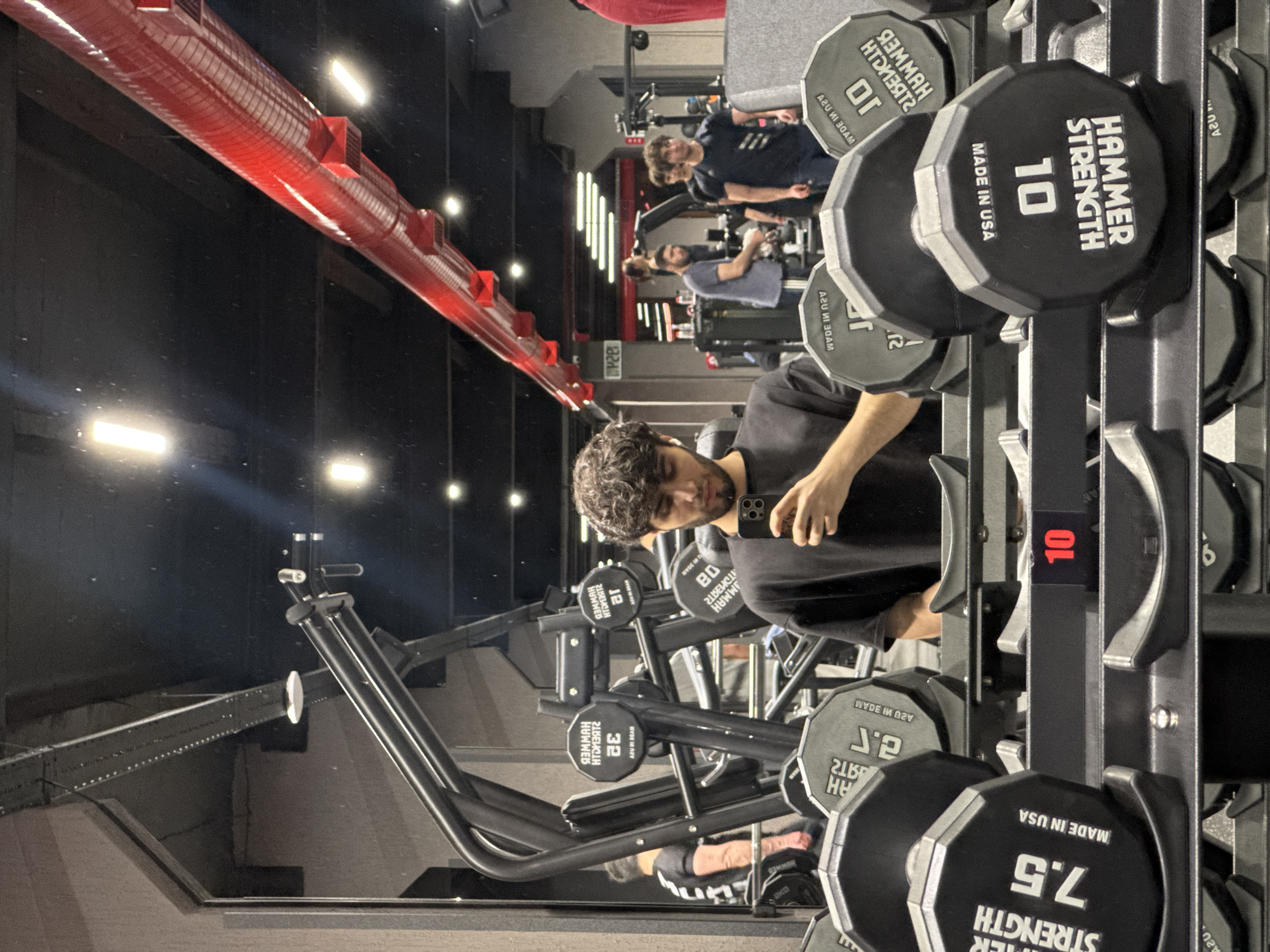 A person is visible in a gym setting, looking at a mobile phone, which they hold in their right hand. The person has curly hair. The person appears to be a reflection in a mirror. In the foreground are racks of dumbbells with "Hammer Strength" branding and weight indications in pounds. Behind the person in the mirror are other people, also in the gym. The gym has black walls and a red ventilation system on the ceiling. The scene appears to be indoors. The location appears to be the Elite Plaza Business Center in Yerevan, Armenia, based on matching architectural features.