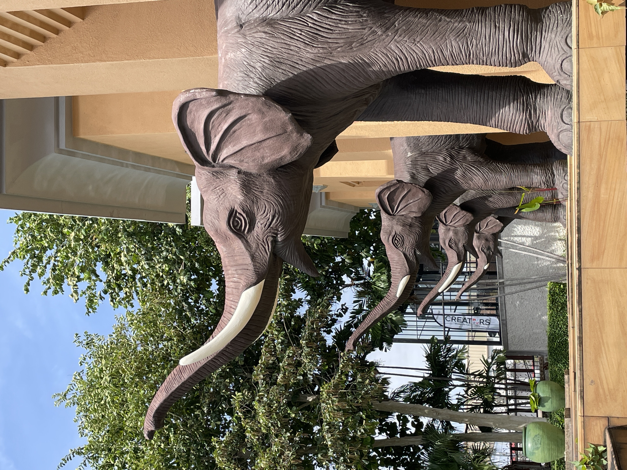 The image shows a line of large elephant statues in front of a building. The statues are brown and appear to be made of concrete or a similar material. They are arranged in a procession, with the largest elephant in the foreground and smaller elephants behind it. The building behind the statues is painted a light orange and has a modern architectural style. There are trees and foliage in the background, suggesting an outdoor setting. The sky is blue and clear, indicating a sunny day. The scene is likely located in Nong Prue, Thailand, possibly a public area or a commercial establishment. No people are visible in the image.