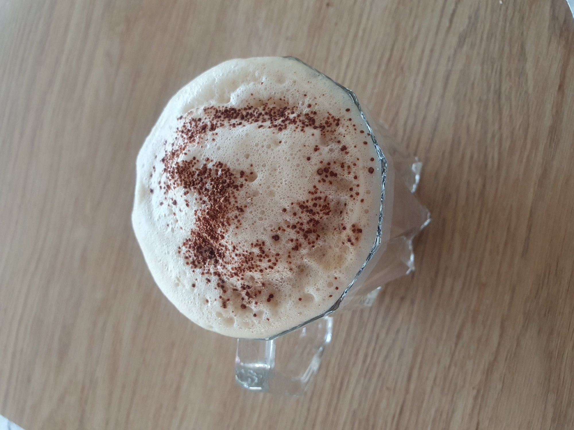 A close-up, high-angle shot shows a glass mug filled with a frothy beverage, possibly coffee or a latte, topped with a dusting of cocoa powder arranged in a pattern resembling the letter 'M'. The mug is transparent with a glass handle and a metallic rim around the top. It rests on a light brown wooden surface. The background is blurred, suggesting the image was taken indoors, potentially in a cafe or home setting. There is no visible indication of the city of Kheyrabad, Iran, or any specific geographical markers.