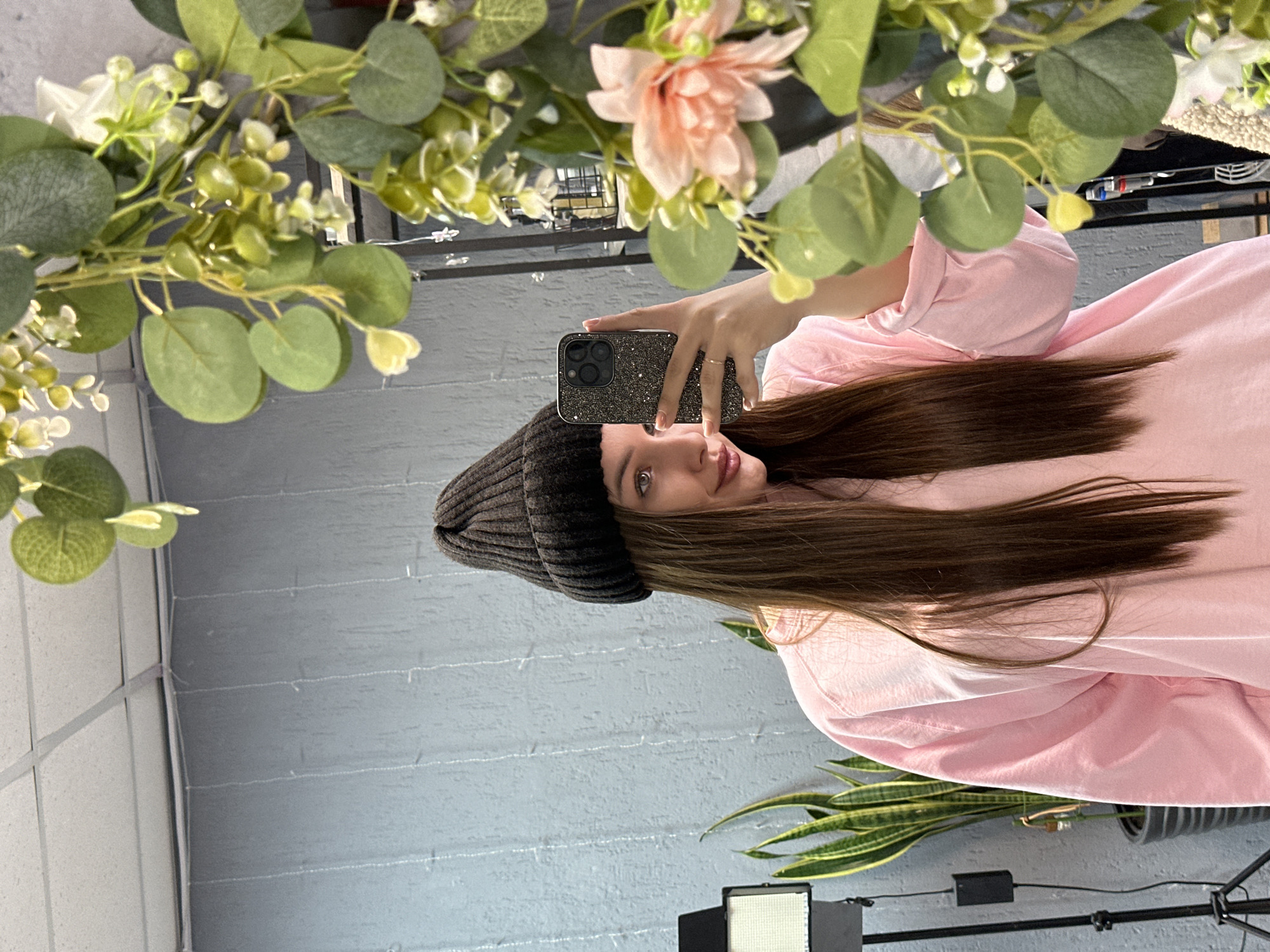 A female subject is depicted in a mirror selfie taken indoors in Chaikovsky, Russia. The subject, a young adult with long, straight brown hair, wears a black ribbed beanie and a loose-fitting pink long-sleeved shirt. She holds a smartphone, encased in a dark, sparkly protective cover, in her left hand, which is raised to capture her reflection. A simple ring is visible on her left ring finger, and her fingernails are short and light-colored. Her visible eye is light-colored, and she exhibits a slight smile.
The right foreground of the image is partially obscured by a decorative artificial garland featuring green leaves, small white blossoms, and a prominent pale pink flower. In the background of the reflection, a light grey textured wall with subtle vertical striations is visible. Above the wall, a white suspended ceiling with grid tiles can be seen. To the left of the subject's reflection, a tall potted Snake Plant (Sansevieria trifasciata) with green and yellow variegated leaves stands in a dark, ribbed pot. Adjacent to the plant is a black professional light stand with a rectangular white panel light attached. The far right background of the reflection, behind the garland, shows indistinct reflections, possibly of shelving or clothing racks.