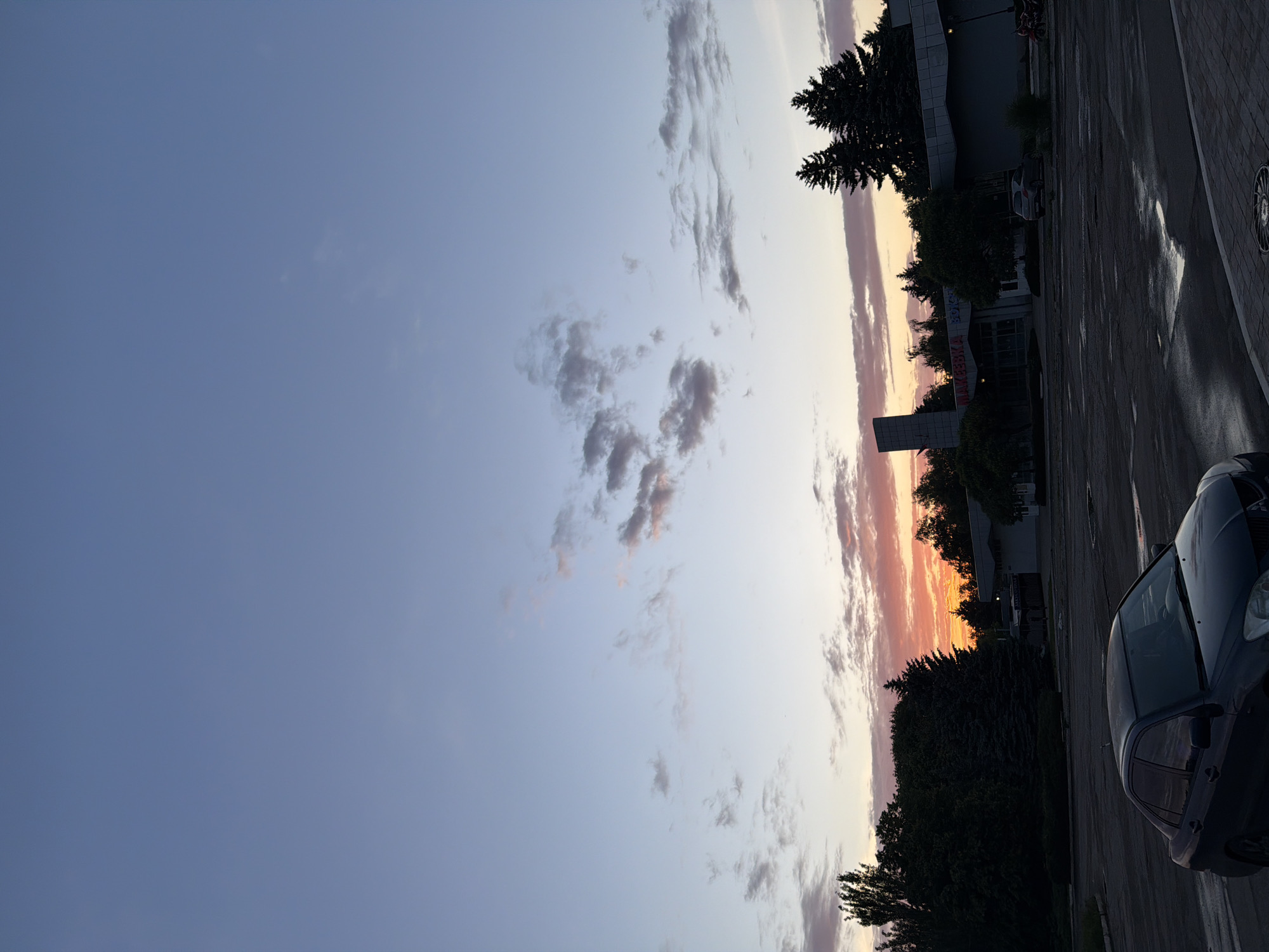 The image depicts an exterior scene at dusk, featuring a wide expanse of sky transitioning from a deep blue at the top to softer pastel shades near the horizon. Scattered clouds are visible, some tinged with pink and orange from the setting sun. In the middle ground, a building with a prominent sign displaying Cyrillic lettering is partially obscured by trees. The building appears to be a commercial establishment, possibly a supermarket, given its size and the architectural style. A dark-colored sedan is parked in the foreground, facing away from the viewer, on a wet asphalt surface that reflects the twilight sky. Several other vehicles and a motorcycle are visible in the background, suggesting a parking lot or an open area near a commercial zone. The overall atmosphere is serene, capturing the quiet transition from day to night in an urban or suburban setting.