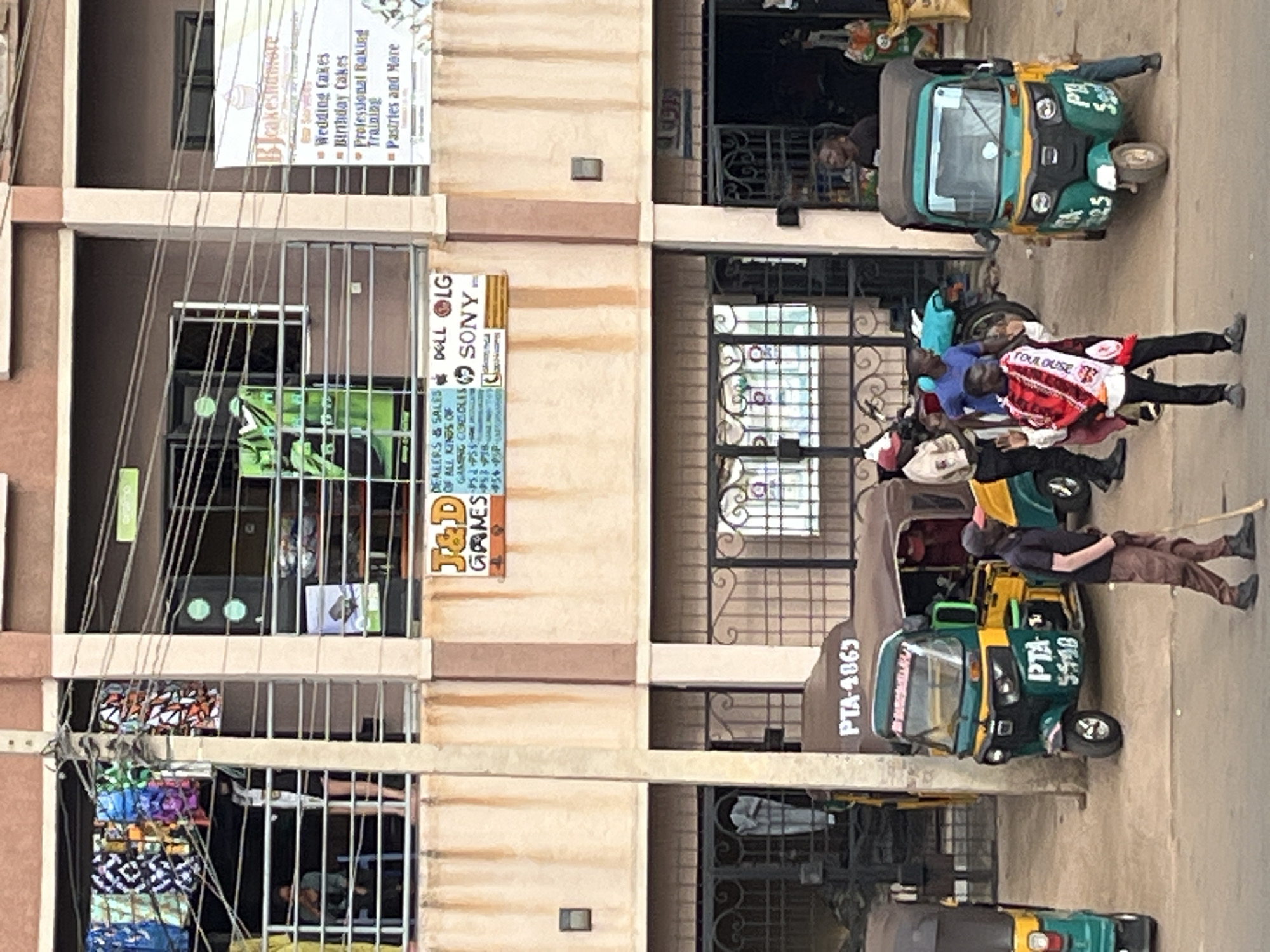 The image depicts a street scene in Bukuru, Nigeria, with a multi-story building as the central focus. On the ground level, there are multiple shops, including "J&D GAMES" which displays signs for Dell, LG, and Sony. Adjacent to this is a bakery named "BJcakesnmore" offering wedding cakes, birthday cakes, and professional baking training. Several auto-rickshaws (Keke Napep) are parked on the street in front of the shops.
The upper levels of the building feature balconies with railings, on which clothing items are displayed for sale. Some individuals are visible on these balconies. On the street, several people are present. One man in the foreground is walking with a stick. Other individuals are interacting near the auto-rickshaws. The overall environment suggests a bustling commercial area.