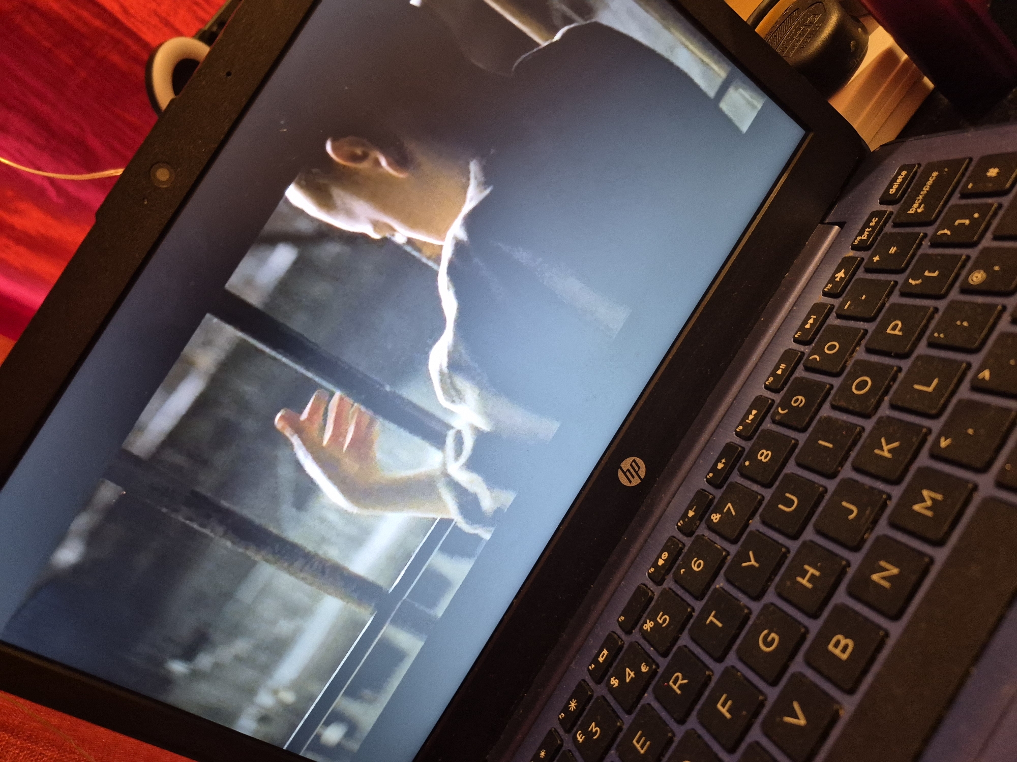 A close-up, angled shot shows a laptop screen displaying a scene from a movie or show. The screen is bright and takes up most of the frame, showing the back of a person's head and shoulders, with their arm raised and hand extended. The background on the screen appears to be a dimly lit interior with bars or grating. The laptop itself is dark blue with a standard QWERTY keyboard visible in the foreground. The HP logo is present on the keyboard deck. The laptop is resting on a surface that is not fully visible, but appears to be a red, textured fabric. No specific location within the city of Sandwell, United Kingdom, can be determined from the image.