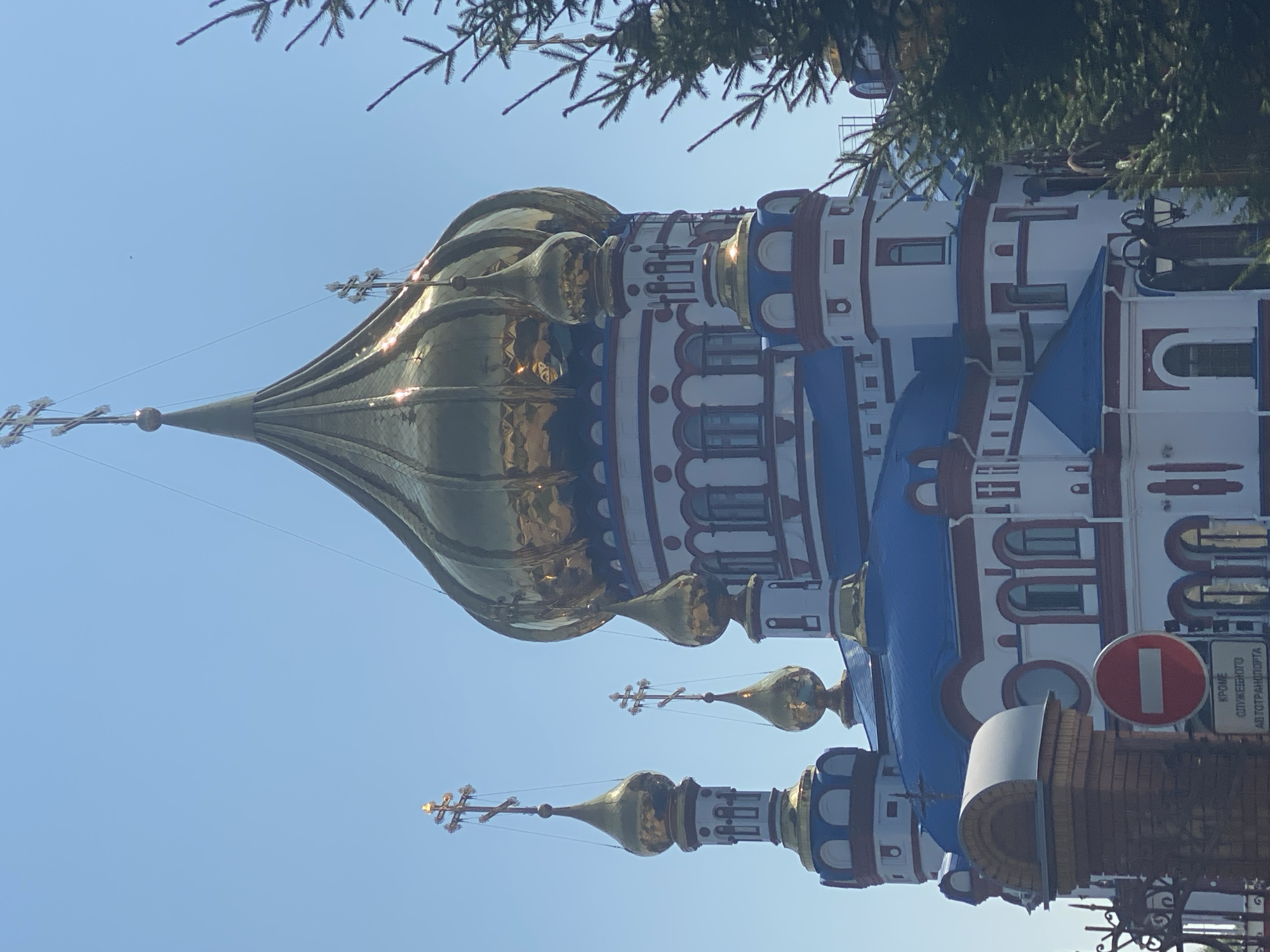 The image shows a close-up view of the golden domes and white facade of the Savior Transfiguration Cathedral in Novokuznetsk, Russia. The cathedral features multiple onion domes, the largest of which is prominent in the center and reflects the sunlight. Smaller domes and crosses are also visible on the spires. The building has white walls with red trim around windows and architectural details, and blue roofs. In the foreground on the left, a brick pillar and a black wrought-iron fence are partially visible, along with a red and white "no entry" sign and a sign with Russian text below it. Evergreen branches from a tree are visible on the right side of the image, partially obscuring the cathedral. The sky is clear and blue.