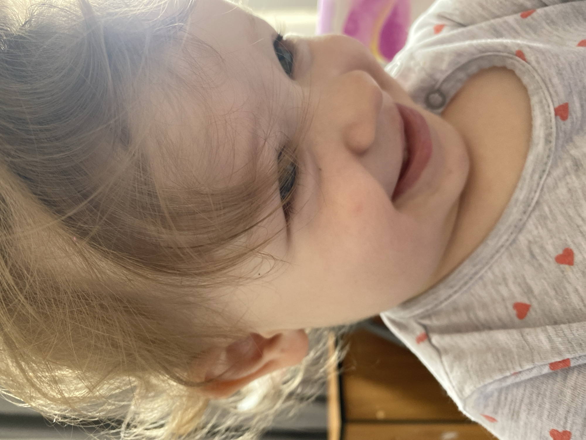 A close-up, slightly tilted view of a young child's face and upper body. The child has fair skin and blonde hair. The child is wearing a gray shirt with small red heart patterns. The child's eyes are partially closed and the mouth is slightly open. A portion of an undefined object is visible in the background.