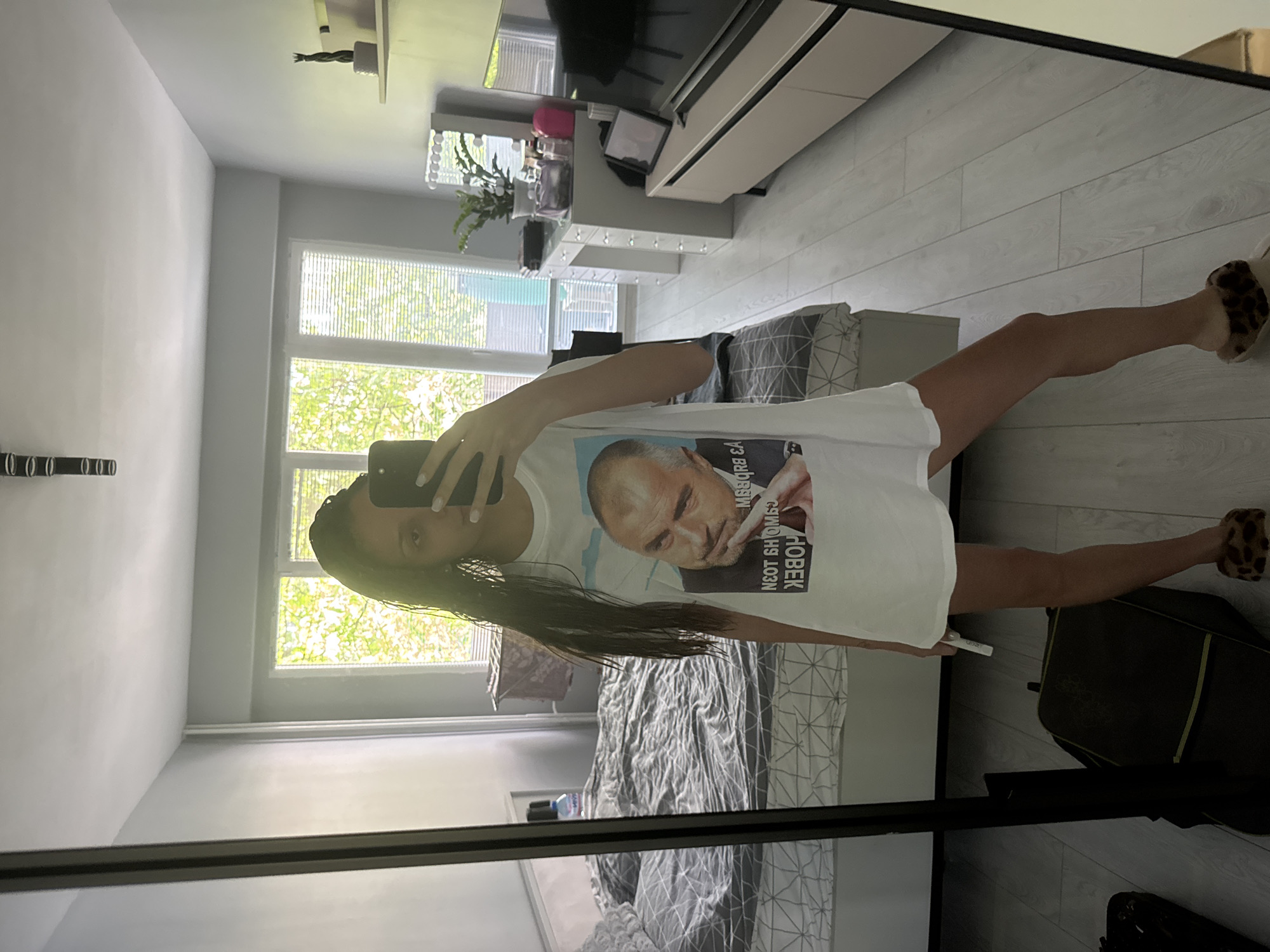 A person is taking a mirror selfie in what appears to be a bedroom. The individual is wearing a white T-shirt with a printed image of a man and text in Cyrillic, along with animal-print slippers. The person is holding a smartphone in their hand, presumably to take the picture.
The bedroom contains a bed with gray bedding, a window with visible trees outside, and a dresser with a TV on top. There is also luggage present.
The image does not contain enough information to reliably identify the exact location in Sofia, Bulgaria.
