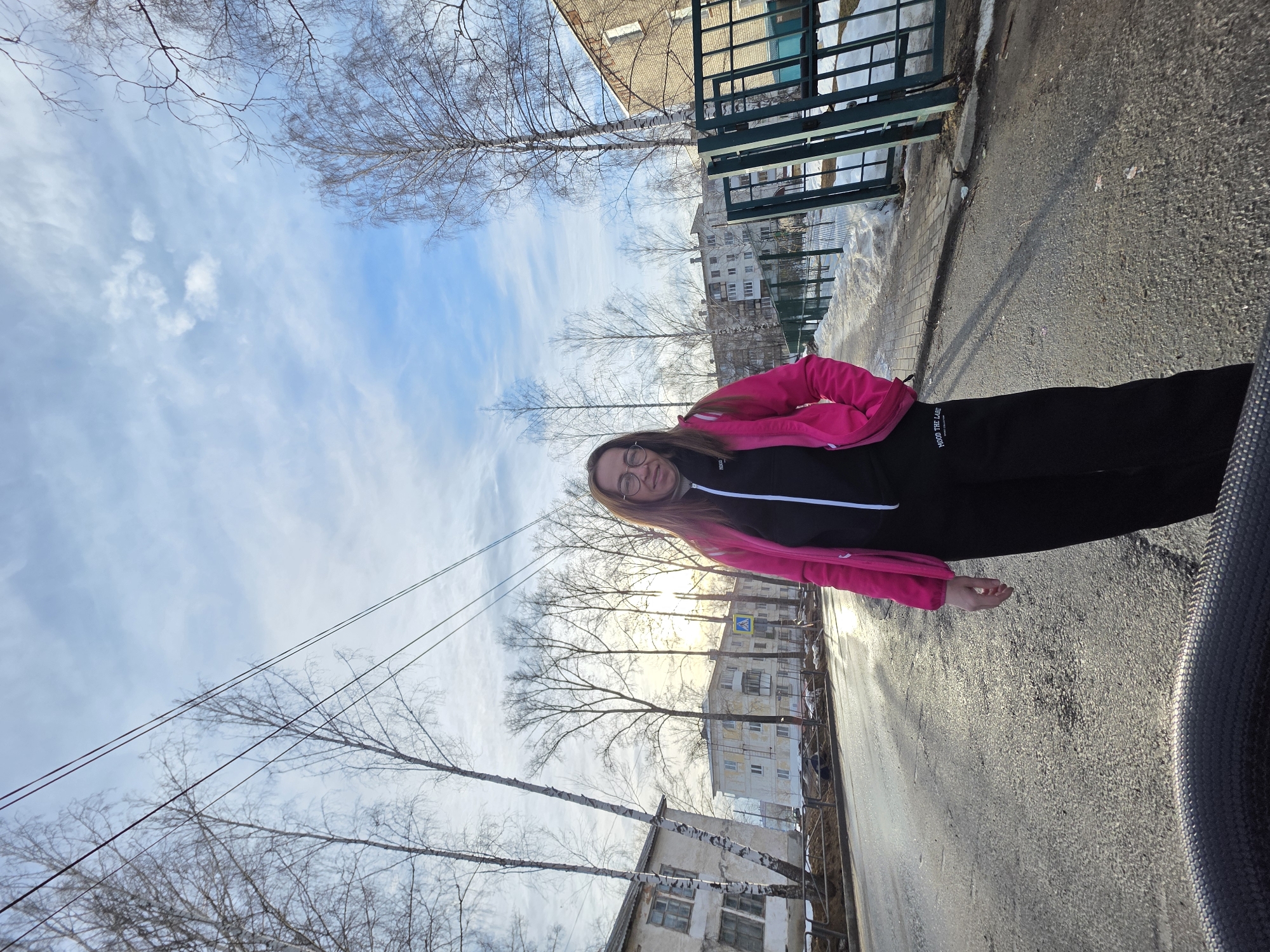 An adult female stands centrally on a wet asphalt street in Chaikovsky, Russia, on a sunny day, likely in late winter or early spring. She wears a bright pink jacket over a black zip-up top and black trousers, looking directly at the camera with a slight smile. Her left hand is in her jacket pocket; her right arm hangs by her side. Bare deciduous trees, including birches, line both sides of the street, which features multi-story residential buildings (apartment blocks) in the background. A light-colored building is visible on the left, and a darker brick building with more apartment blocks behind it is on the right. A dark green metal fence runs along the right side of the street. A blue pedestrian crossing sign with a white triangle and figure is visible in the mid-ground on the left. Patches of snow and slush are present on the sidewalk. Overhead power lines span across a blue sky with scattered white clouds. Low-angle sunlight creates strong reflections on the wet road surface. A dark, textured circular object is partially visible at the bottom center of the frame.