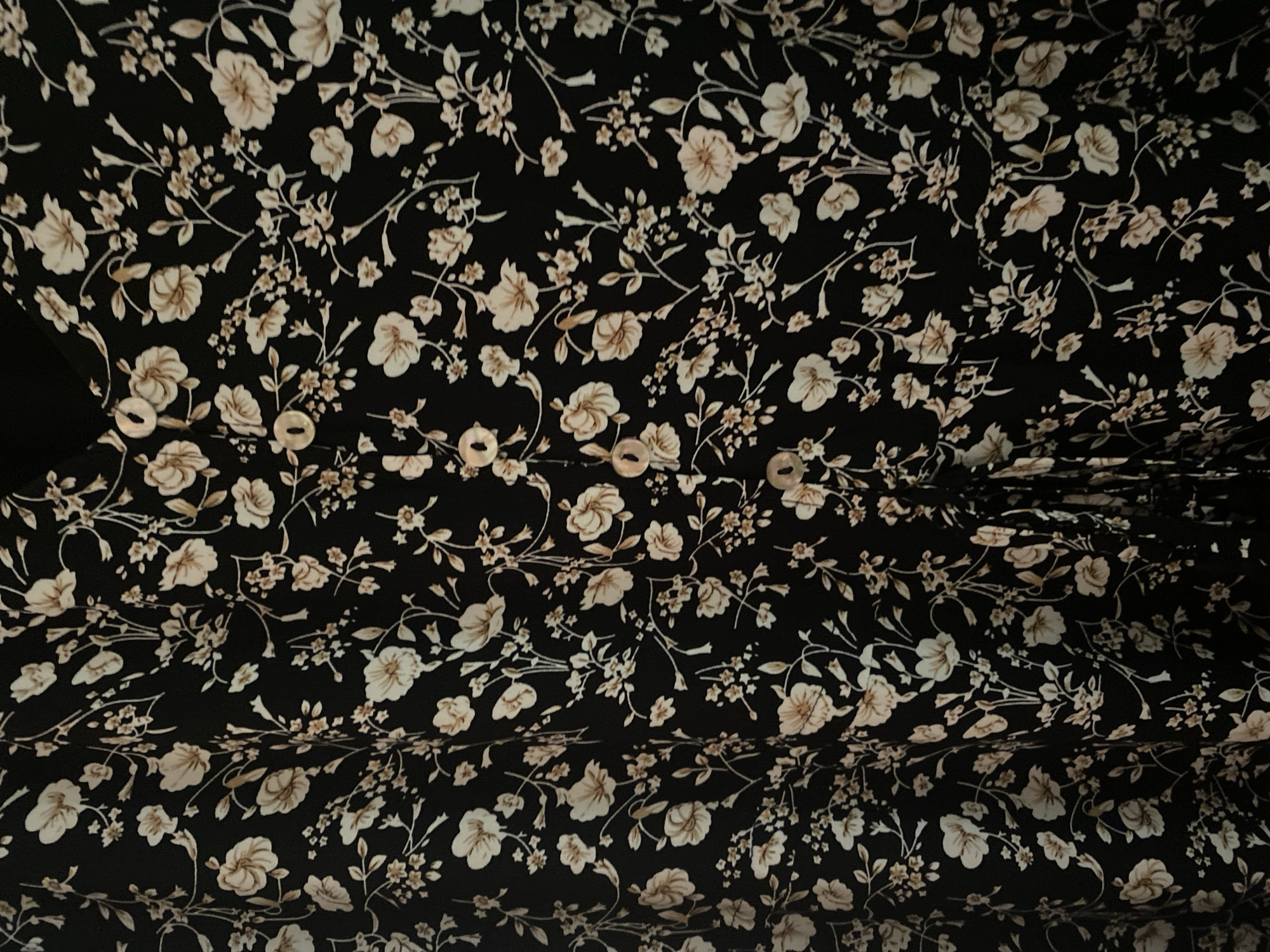 The image shows a close-up of a black fabric with a floral print. The print consists of small, light-colored flowers and leaves scattered across the black background. There are also several pearl-like buttons visible on the fabric, suggesting it is part of a garment. The lighting is somewhat dim, casting subtle shadows that highlight the texture of the fabric. The scene is devoid of any specific urban or natural landmarks, making it impossible to determine its location within Chernogorsk, Russia, or any other city. The focus is solely on the textile pattern.