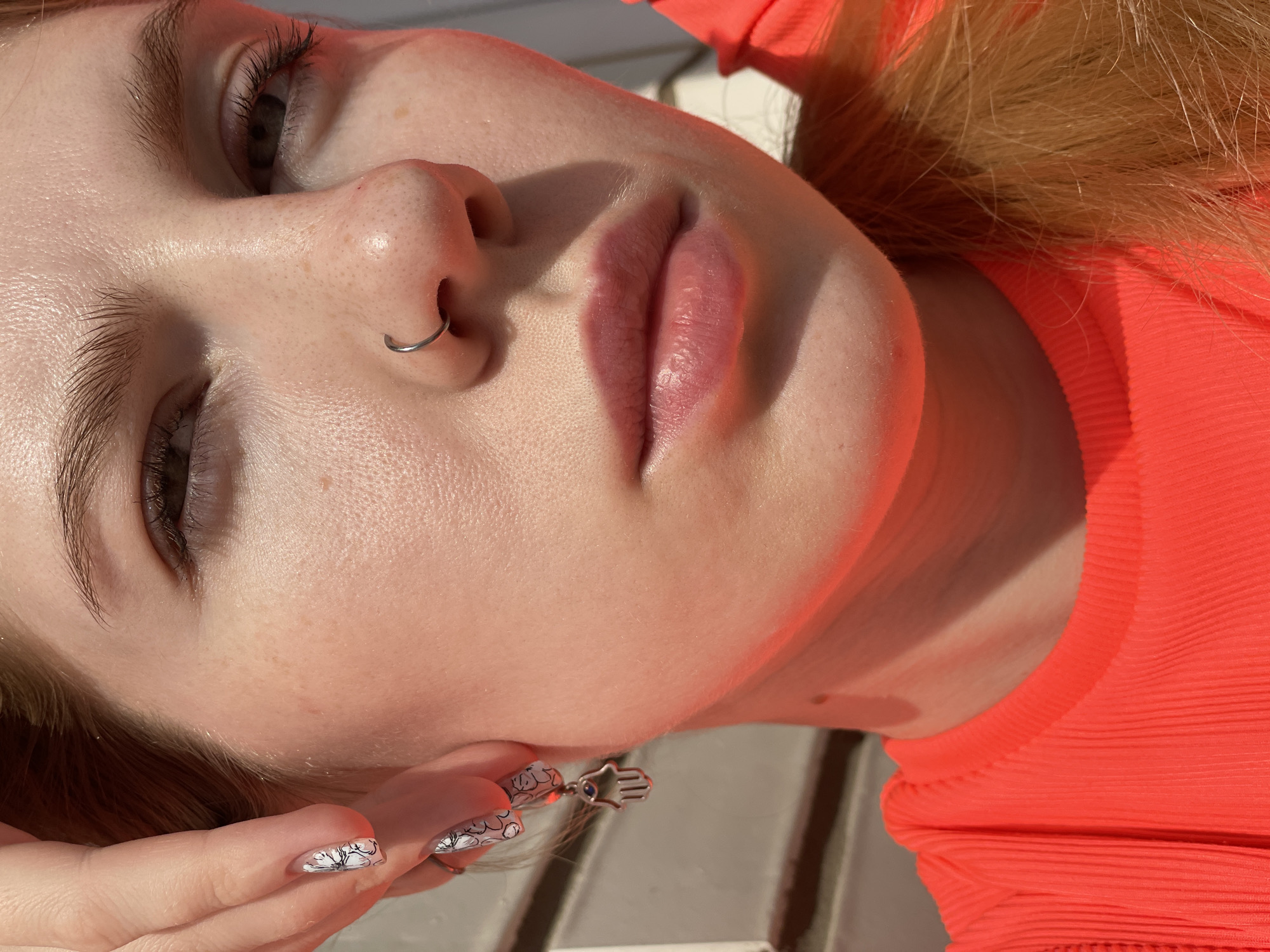 The image is a close-up, upward-angled shot of a fair-skinned individual, likely female, with light brown or blonde hair. She is wearing a bright orange top. Her facial features are prominent, including her eyes, nose with a silver nose ring, and full lips. She is wearing manicured nails with a black and white floral design on her nails. She is wearing a silver earring shaped like a hamsa hand. The lighting appears to be natural sunlight. There is no indication of Izhevsk, Russia, in the image.