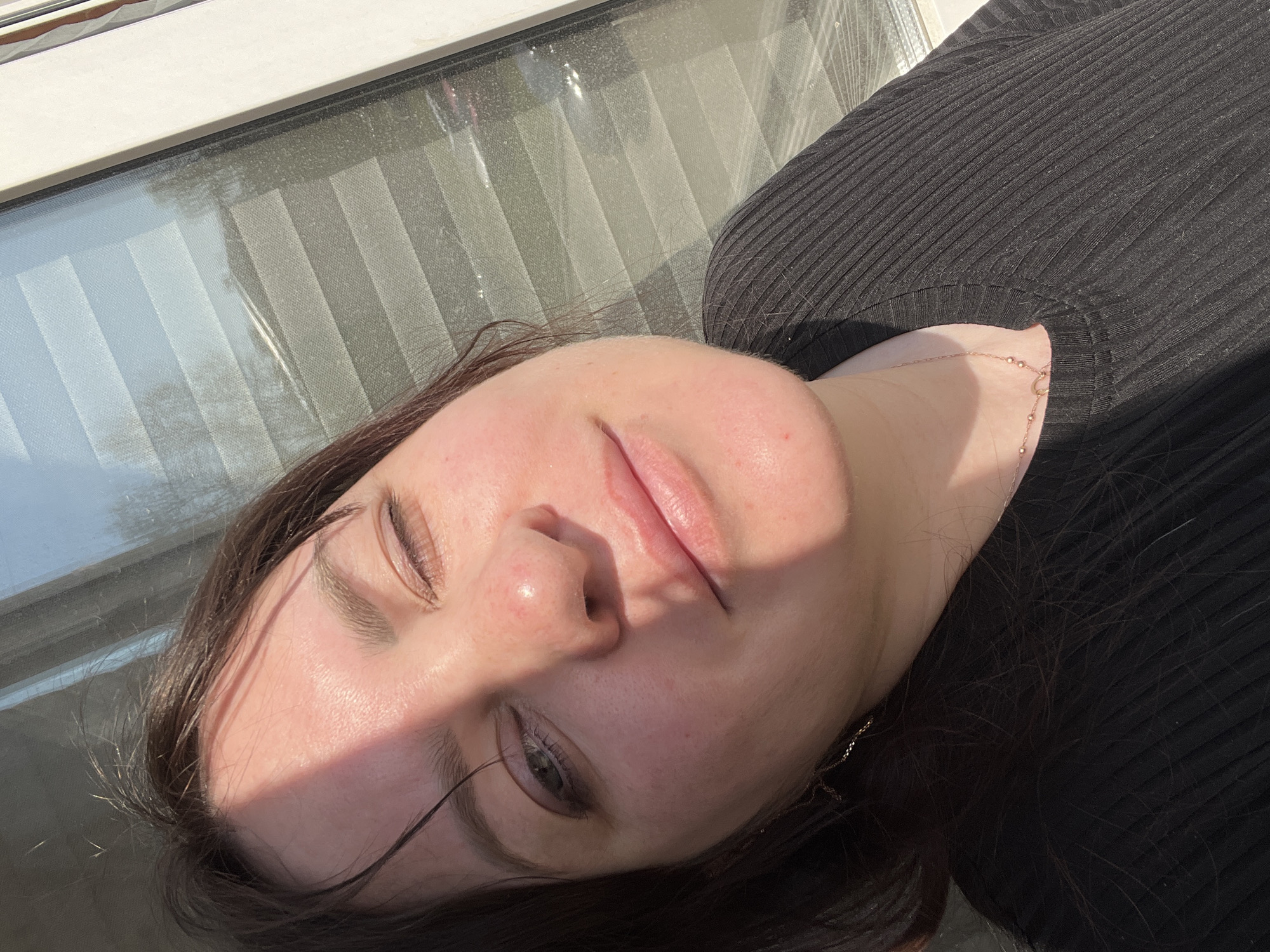 A young woman with dark brown hair is lying down indoors next to a window with drawn blinds. She is wearing a black ribbed shirt. Her eyes are partially closed, and she is facing upwards toward the light. A gold necklace is visible around her neck. The location is unknown and cannot be determined from the image. The image contains no information regarding the city of Noviy, Russia.