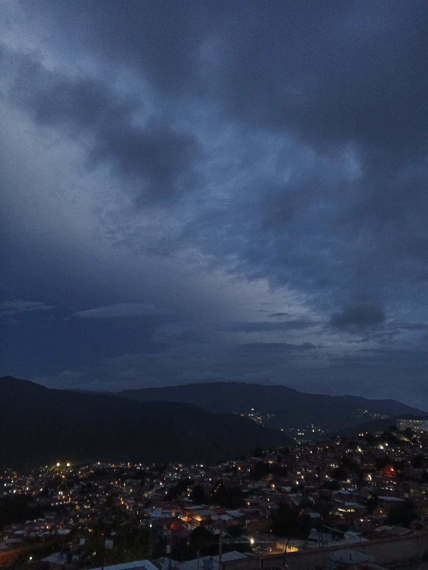 The image depicts a panoramic view of a densely populated hillside neighborhood in Caracas, Venezuela, at dusk or night. The sky is filled with dark, dramatic clouds, suggesting an impending storm or the aftermath of one. Below the sky, silhouetted mountains form the backdrop to the sprawling city.
The foreground and middle ground are dominated by a hillside covered with numerous buildings. Many of these buildings are illuminated by warm, yellow lights, indicating that it is nighttime and residents are inside their homes. The density of the settlement is evident, with houses packed closely together, following the contours of the terrain. The architecture appears to be a mix of residential structures, with various roof types visible, including red tiles and flatter surfaces.
In the distance, on the slopes of the mountains, scattered lights can be seen, suggesting settlements or structures further up the hills. The overall impression is one of a vibrant, active community nestled within a challenging, mountainous landscape. There are no visible individuals or specific actions taking place, but the presence of lights implies human activity and daily life within the settlement. The environment is urban, characterized by the dense housing and infrastructure, set against a natural mountainous backdrop. The lighting is a key element, with the contrast between the dark sky and the illuminated city creating a dramatic and atmospheric scene.
