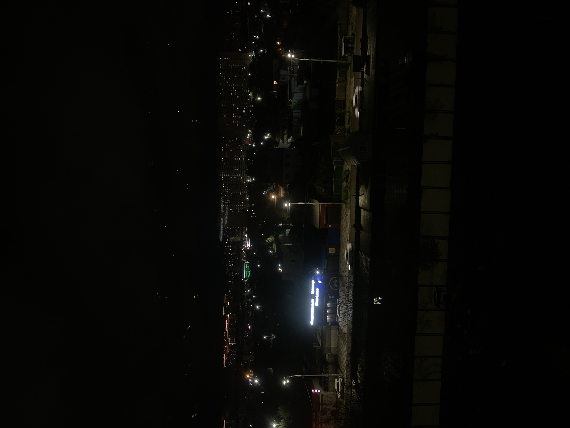 Here is a detailed description of the image:
The image shows a nighttime view of a city, likely taken from a higher vantage point such as a building or hill. On the right side of the frame, there is a dark, silhouetted building, possibly residential, with visible rows of windows. Adjacent to this building is what appears to be a yard or open space, also largely in shadow.
Moving towards the left, the cityscape opens up. The buildings in the city are illuminated, with various lights creating a dynamic pattern against the night sky. There are signs of street lighting, commercial signs, and building lights, indicating urban activity. One lit-up sign stands out with a bright horizontal line and some vertical elements, likely a business or establishment name.
The environment is dark, with the light sources providing the main contrast. The lack of specific landmarks makes it challenging to pinpoint the exact location within the city. There are no visible people or overt actions taking place in the frame. The overall interaction is that of an urban setting at night, characterized by the presence of buildings, lights, and infrastructure.