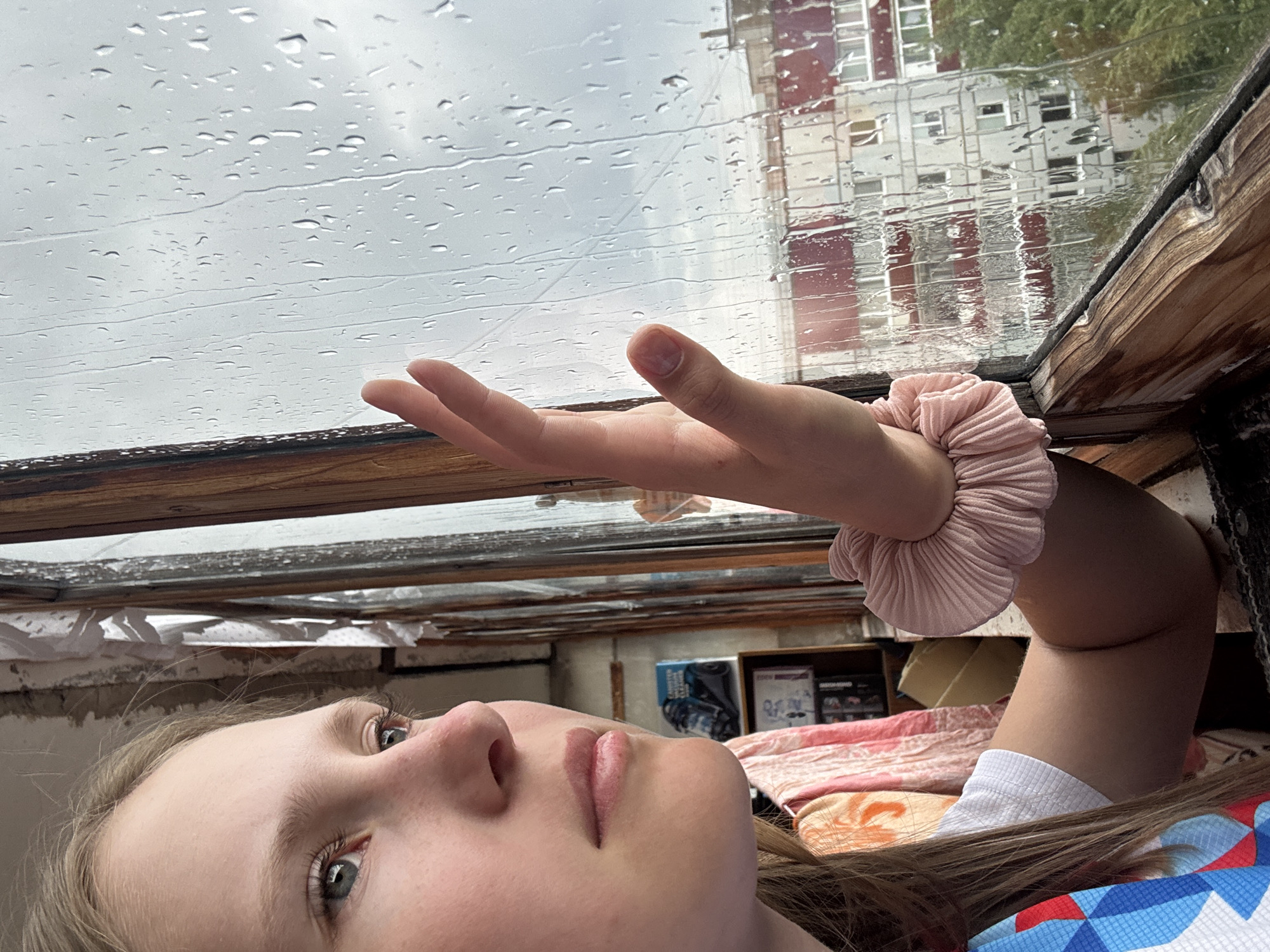 The image depicts a person lying on their back looking up through a rain-streaked window. The person has fair skin, blue eyes, and light brown hair. They are wearing a pink scrunchie on their arm and a garment with a geometric blue and red pattern. The window has a wooden frame and is covered in raindrops. Reflected in the window is a building with red and white facade. In the background, behind the person, are various objects including a blanket, boxes, and shelves. Given the location provided, the scene is likely taken in an interior space in Chaikovsky, Russia with a view of a building across the street, on a rainy day.