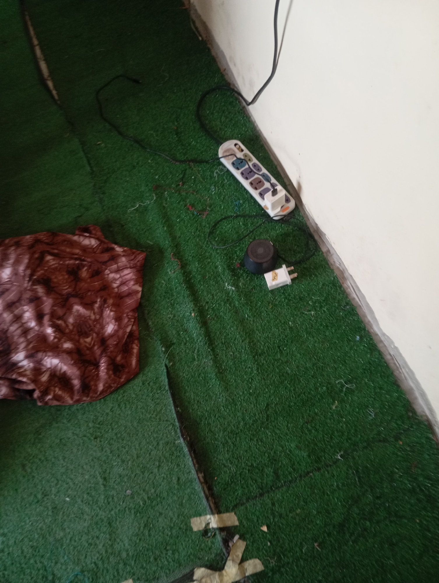 An overhead shot shows a section of a room with an artificial grass floor. In the upper right corner, a white electrical power strip is plugged into an outlet, with several plugs inserted into it. Next to the power strip, a small black speaker sits on the floor. A white plug is also on the floor near the speaker. In the upper left quadrant of the image, a folded piece of dark brown fabric with a marbled pattern lies on the grass. The wall in the background is plain white.