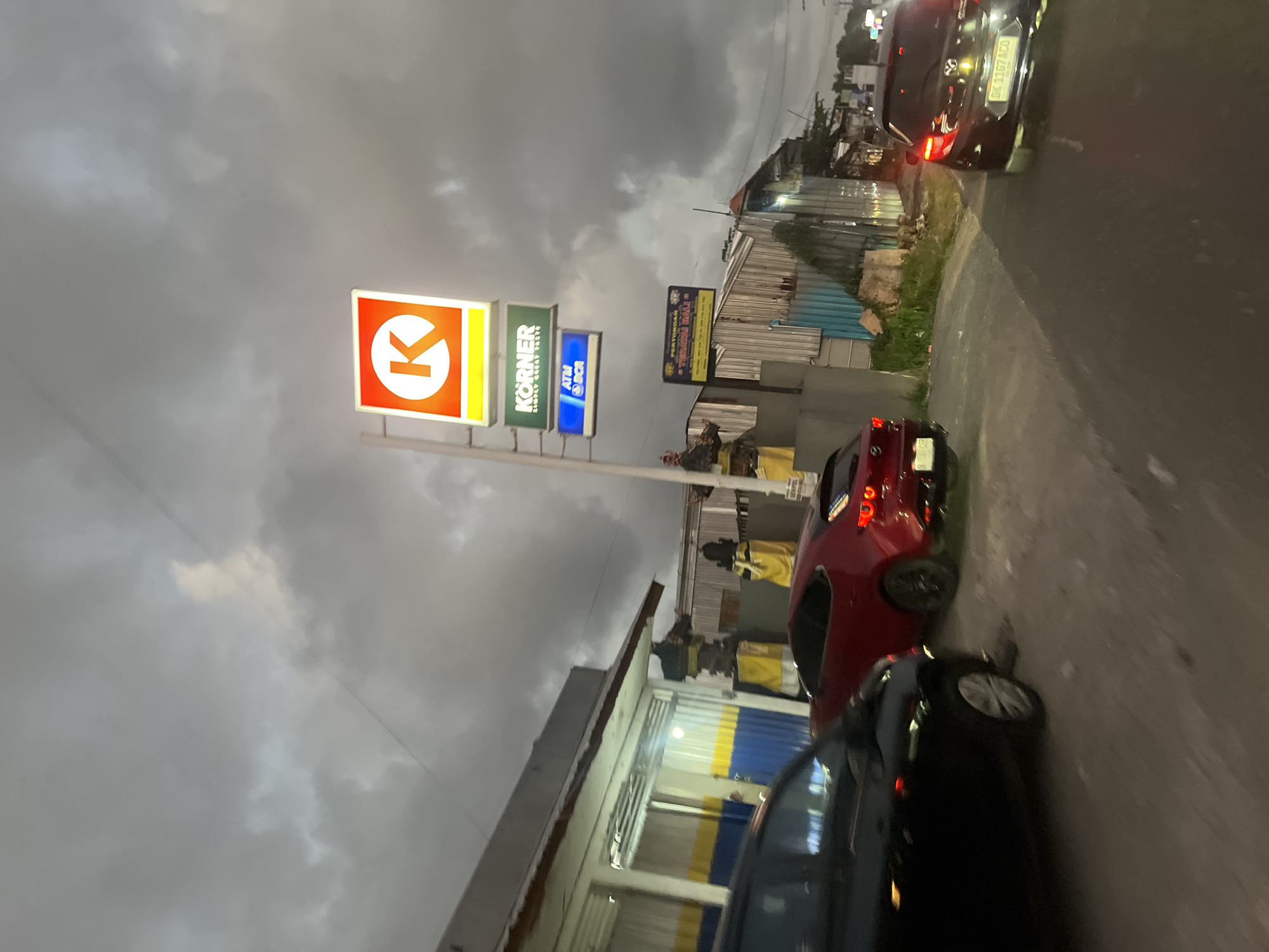 The image shows a street scene at dusk in Mengwi, Indonesia, under a cloudy sky. A white pole with stacked signs stands prominently on the right side of the road. The top sign is illuminated with a red square and an orange "K" logo. Below it is a green rectangular sign reading "KÖRNER SIMPLY GREAT TASTE" and a blue sign labeled "ATM BCA".
In the foreground, a black car is parked on the left, and a red car is positioned behind it. Further down the road, a dark-colored vehicle with its brake lights illuminated is visible. To the left of the signs, a building with a white facade, blue and yellow striped siding, and an open doorway is partially visible. Several decorative statues are present near the building. A smaller yellow sign is affixed to a building in the background. The street appears to be paved, with vegetation growing along the side. The overall atmosphere is dim, suggesting the end of the day or overcast conditions.