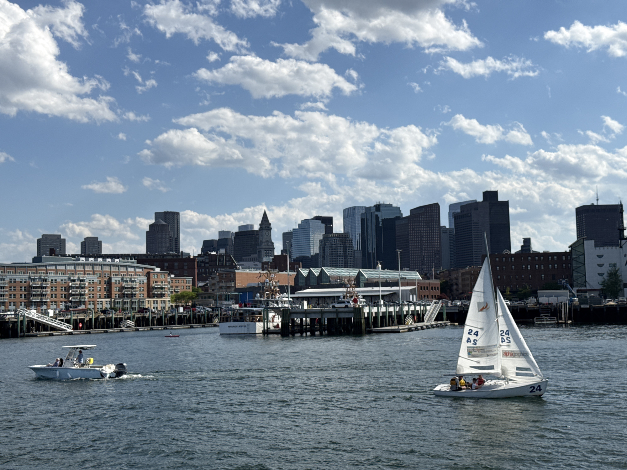 The image captures a waterfront scene in Boston, Massachusetts. In the foreground, a small sailboat with three visible occupants is sailing on the water. To the left, a powerboat with two people aboard is also moving through the water. In the midground, there are docks and buildings with vehicles and some boats, including a vessel named "Maurice Jester". The background is dominated by the Boston skyline with various skyscrapers and buildings, under a sky with scattered cumulus clouds. The scene appears to be taken during a sunny day.