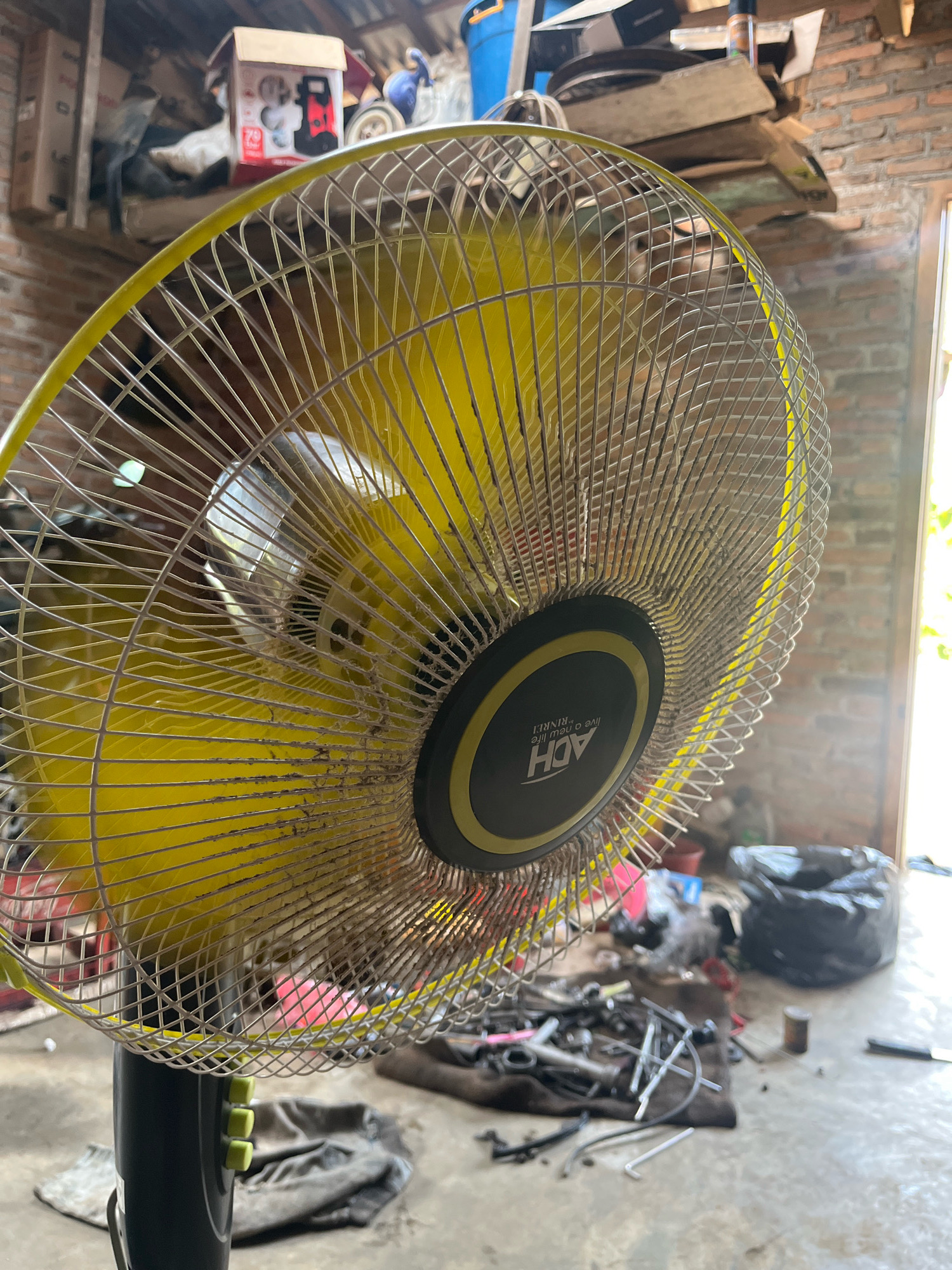 The image depicts a yellow electric fan positioned in the foreground, angled towards the right. The fan has a metallic grille with a yellow rim and a central black circular panel featuring white text. Behind the fan, the environment appears to be an indoor workshop or storage area. The background includes a brick wall, shelves laden with various items, a blue plastic container, and a black trash bag. Scattered on the floor are tools and what appears to be automotive parts. The lighting suggests an indoor setting, possibly with natural light entering from a doorway or window on the right. The overall scene conveys a sense of a working or storage space that is somewhat cluttered. The location is identified as Kebumen, Indonesia.