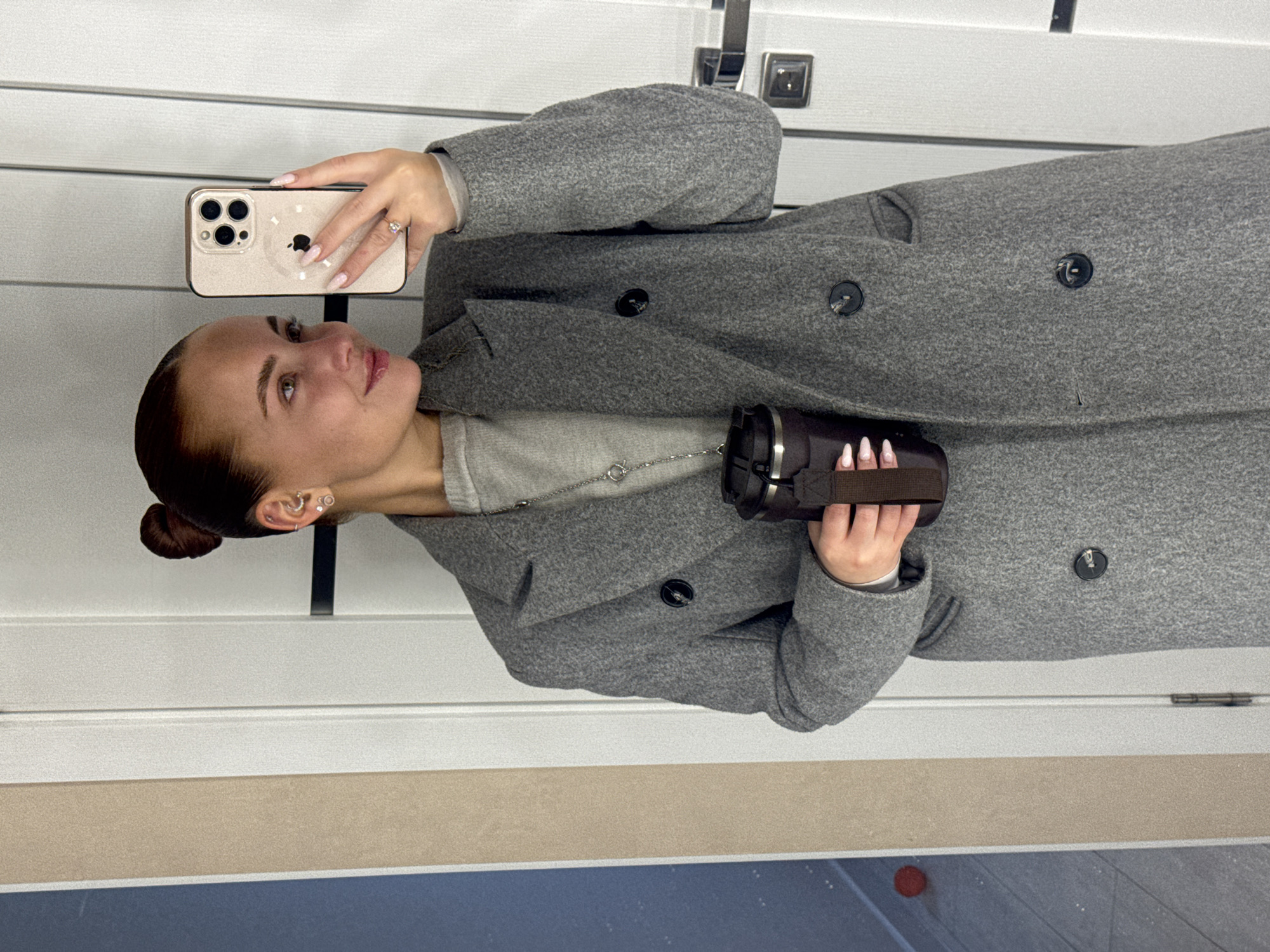 A young woman is captured in what appears to be a mirror selfie, oriented vertically in the original image but described as if upright. She has her dark hair pulled back into a neat bun and wears multiple small hoop earrings. She is dressed in a light gray long-sleeved top, adorned with a delicate silver chain necklace, and a chunky, double-breasted grey wool coat with large dark buttons.
In her right hand, she holds a large iPhone, likely a Pro Max model, in a light, possibly champagne-colored, case that features a cutout of the Apple logo on the back. Her left hand holds a dark-colored insulated bottle or cup, secured with a brown strap. Her nails are manicured with a light pink or nude polish. She wears a ring with a clear stone on her right ring finger. Her gaze is directed slightly upwards and to her left, with a subtle, content smile.
The background consists of a white door with horizontal paneling and visible dark hardware (handles/hinges). Below the door, there's a light beige stripe, transitioning to a darker, possibly tiled, floor area. A small red circular object is visible on the floor in the lower right corner. The scene appears to be indoors. No explicit text is visible beyond the Apple logo on the phone case. The location context provided is Chaikovsky, Russia, though no specific visual cues confirm this.