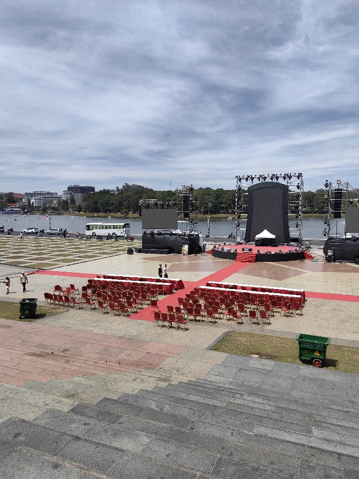 A stage with a large screen is set up on a paved plaza near a body of water. Red carpets lead to the stage, lined with rows of red chairs. Structures resembling lighting rigs flank the screen. A bus and some vehicles are parked nearby. The foreground features a set of wide, gray concrete steps leading down to the plaza. Two green garbage bins are visible on the plaza.