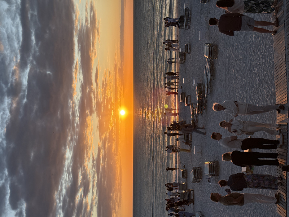 The image depicts a beach scene at sunset in Pelican Bay, United States of America. A bright, circular orange sun is low on the horizon, positioned centrally over a calm body of water, which reflects the sun's light as a golden path. The sky above features scattered dark grey and orange-toned clouds. The sandy beach is populated by approximately 50 to 100 individuals, organized in various small groups. A concentration of people stands near the water's edge, many appearing as silhouettes against the setting sun. Further inland on the sand, numerous beach lounge chairs are arranged in rows, some stacked, alongside several small white tables. In the immediate foreground, a wooden slatted platform extends, where a group of six individuals stands, engaged in conversation. The scene is illuminated by the natural light of the setting sun, casting a warm glow and long shadows.