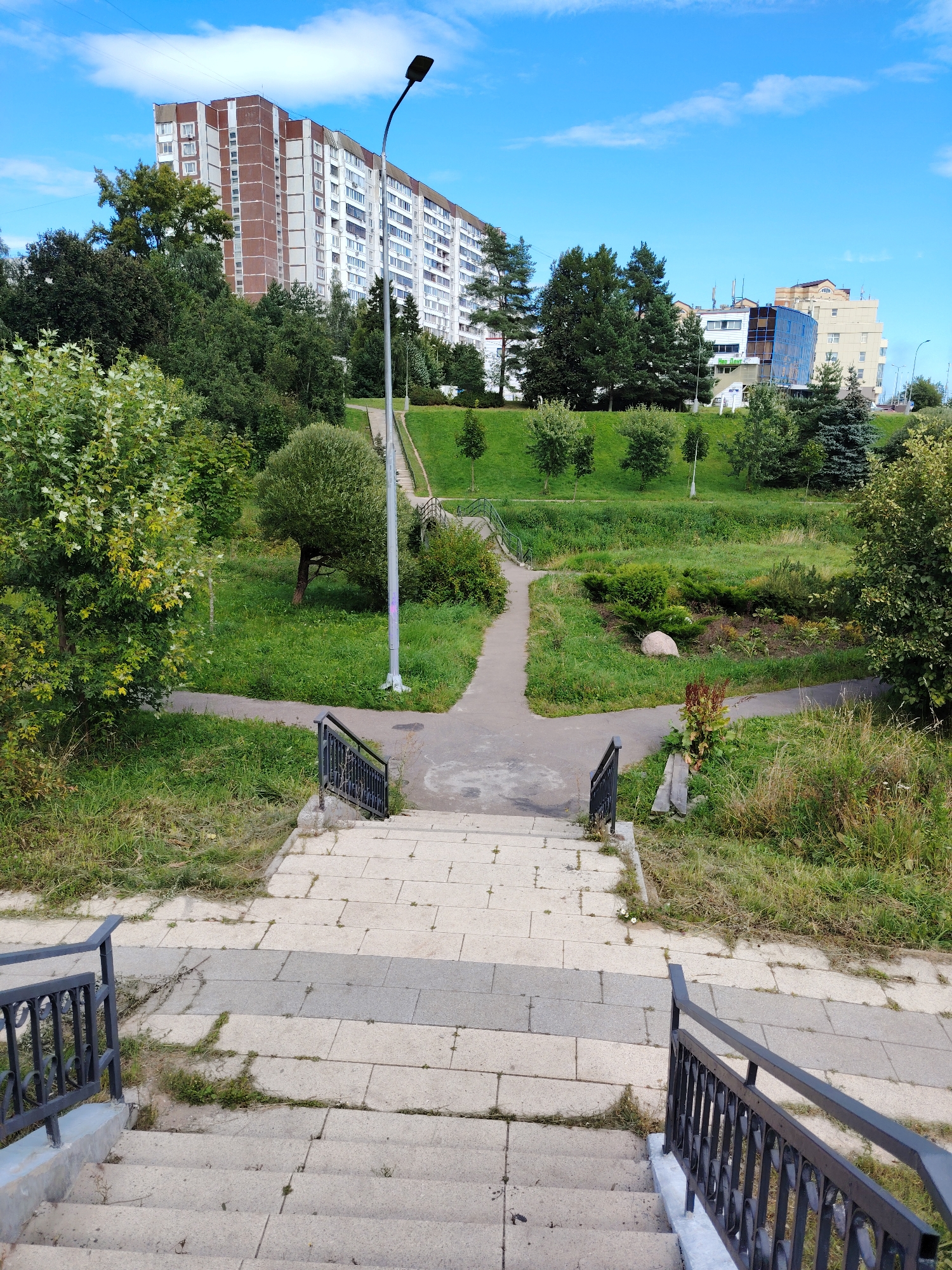 A wide shot captures a sunny day in Zelenograd, Russia, showcasing a multi-level park or public space. In the foreground, a stone staircase with black metal railings descends, bordered by patches of grass and weeds. The path then transitions to a paved walkway that forks, leading in two directions. To the left, a lush green tree with light-colored leaves stands prominently.
Beyond the walkway, the terrain rises, covered in manicured green grass and dotted with various trees and shrubs. A few smaller trees are scattered across the slope. A single street lamp on a tall pole is positioned on the right side of the ascending path.
In the mid-ground, a picturesque scene unfolds with a small bridge arching over a vegetated area. Further into the distance, a large, pale boulder rests within a planted garden bed.
The background is dominated by a large, multi-story residential building with a reddish-brown facade, featuring numerous windows. To its right, another modern building with a blue glass facade and white accents is visible. The sky above is a bright blue, adorned with scattered white clouds. The overall impression is one of a well-maintained urban green space.