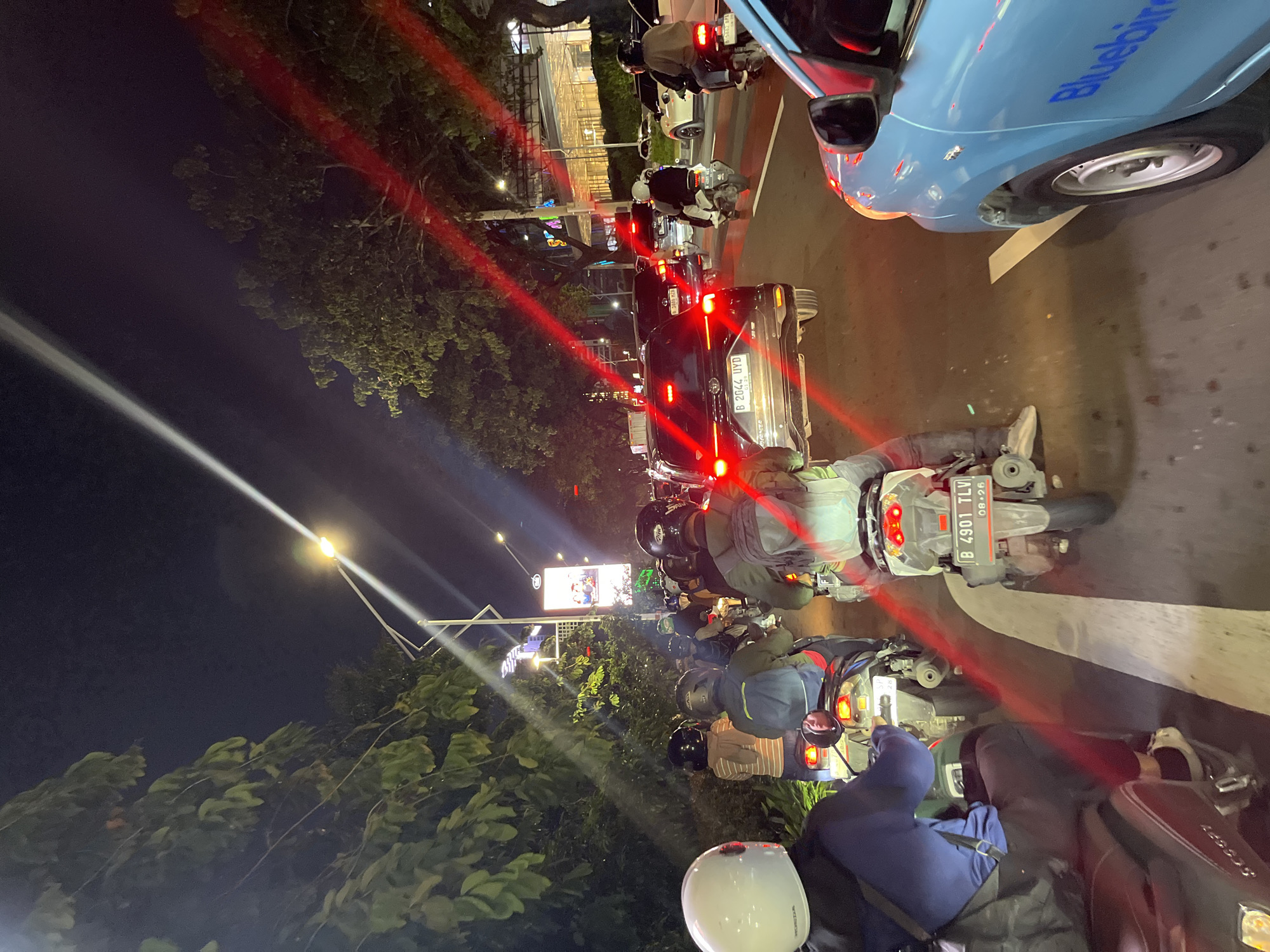The image captures a bustling night scene of heavy traffic on a multi-lane street in Central Jakarta, Indonesia. The perspective is from a motorbike rider, looking forward and slightly upwards into the congested roadway. The time of day is clearly night, evidenced by the dark sky, illuminated streetlights, and prominent vehicle lights.
Numerous motorbikes, mostly scooters, dominate the foreground and mid-ground, with many riders and passengers wearing helmets. Cars are also present, including a light blue Bluebird taxi partially visible on the upper right side and a dark SUV/MPV in the middle distance. Red streaks of light, likely from vehicle tail lights, traverse the frame, suggesting slow movement or stationary traffic and potentially a longer exposure or camera motion. A bright white light streak from a streetlight also cuts across the scene.
Notable details include the characteristic dense motorbike traffic common in Indonesian cities. Streetlights illuminate parts of the road and the lush green trees lining the street. In the background, illuminated buildings hint at an urban environment.
Visible text includes:
- On the blue car, "Bluebird" is partially visible.
- A motorbike license plate reads "B 4901 TLV" with an expiry date "08.26".
- Another car's license plate displays "B 2044 UYD".
- The word "HONDA" is faintly visible on a white helmet in the immediate foreground.
- On the back of the dark SUV/MPV, a faint logo resembling "Avanza" can be discerned.
- On a dark red scooter in the lower right, "KYMCO" is visible.