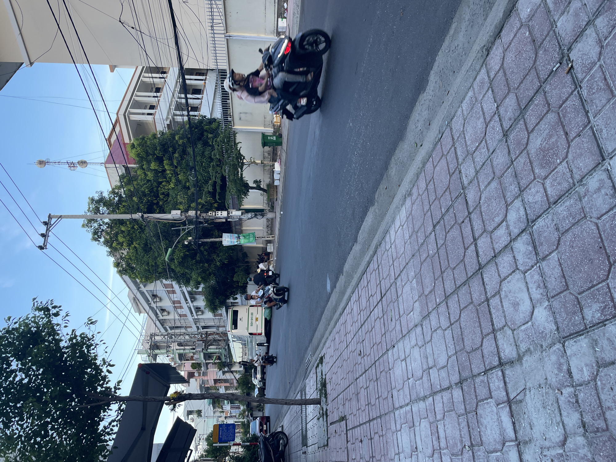 The image depicts a street scene in Nha Trang Ward, Vietnam. A paved sidewalk with red interlocking pavers runs along the left side of the frame, bordered by a street with asphalt paving. Several motorcycles are visible on the street. In the foreground, a black motorcycle with two occupants, one wearing a helmet, is in motion, with its brake lights illuminated. Further down the street, another motorcycle with a single rider is also in motion. A large white bus with the license plate "60H-941.00" is parked or moving slowly on the right side of the street.
Buildings line the right side of the street, exhibiting a mix of architectural styles. One prominent building has a series of white columns and a red-tiled roof, with a telecommunications tower visible on its top. Trees with green foliage are interspersed along the street and sidewalk. Utility poles and overhead power lines crisscross the sky, which is a pale blue with hints of clouds. The scene appears to be captured during the late afternoon or early evening, as indicated by the quality of light. The overall impression is of a typical urban street in Vietnam with active local transportation.