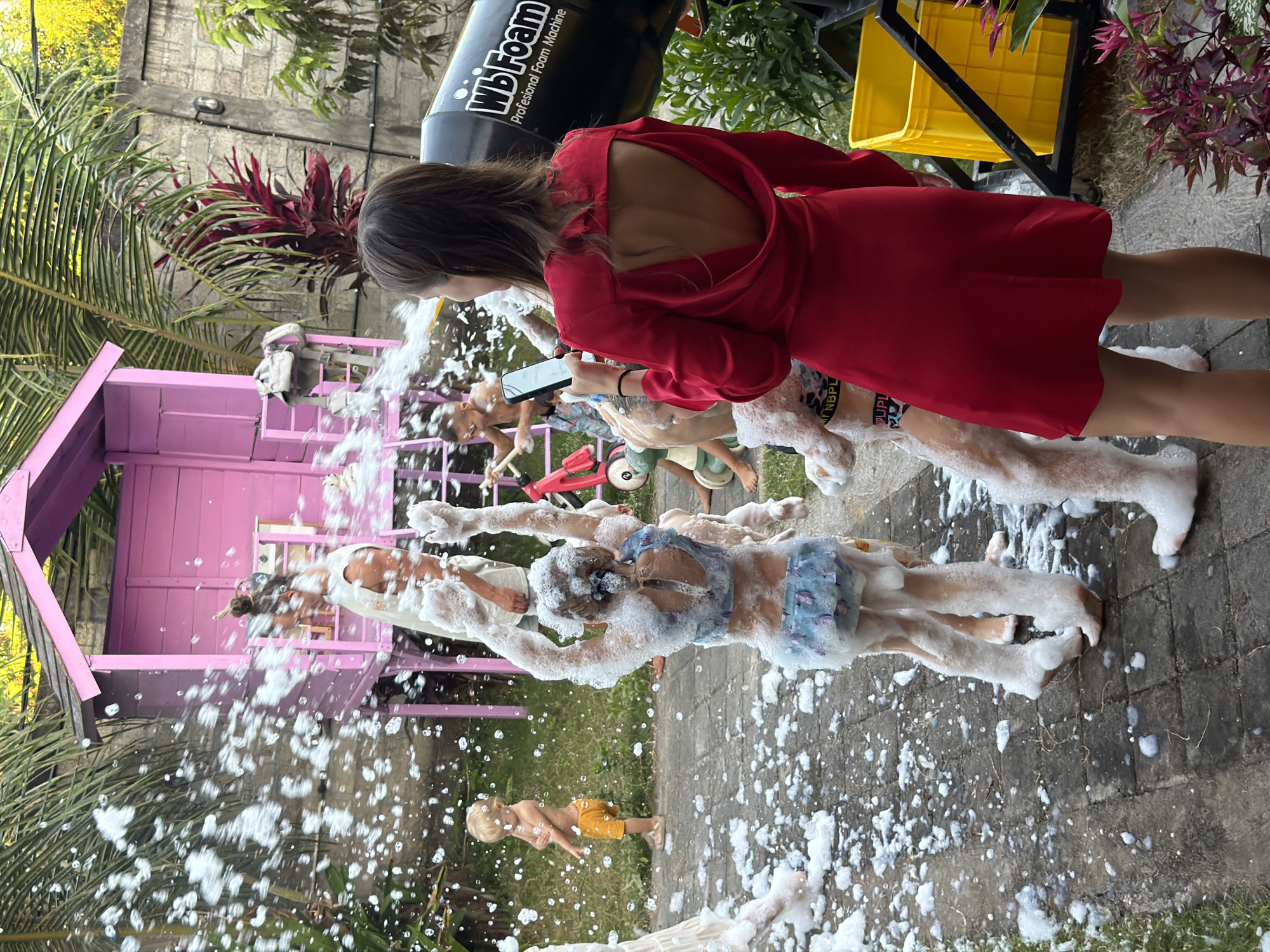 This media file captures a vibrant outdoor foam party in Kuta Selatan, Indonesia. A large black "WbFoam Profesional Foam Machine" is prominent in the upper right, actively dispensing white foam that blankets the scene.
A young woman with long, dark hair, seen from behind, is wearing a striking red dress with an open back. She is holding a smartphone, seemingly capturing the event. Several children are joyfully engaged in the foam. One child, largely covered in foam, is wearing a blue and white patterned swimsuit. Another child in yellow shorts stands near a bright pink wooden playhouse on the left. Other children's arms and legs are visible amidst the bubbly expanse, suggesting a lively gathering.
The setting is an outdoor space with paved ground and patches of grass. Lush green foliage, including palm trees and red-leafed plants, provides a tropical backdrop, consistent with the Kuta Selatan location. Yellow crates are visible near the foam machine. The natural lighting indicates it is daytime. The overall impression is one of playful activity and lighthearted fun. The visible text on the foam machine reads "WbFoam Profesional Foam Machine."