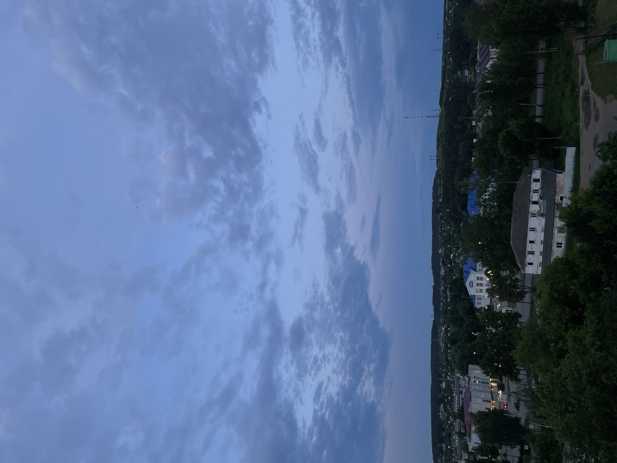 The image captures an aerial view of a town at dusk, with the sky filled with scattered clouds. The scene is oriented vertically, suggesting it was taken from a high vantage point, possibly a building or drone.
The dominant subjects are the landscape and the sky. Below the sky, a town unfolds, characterized by a mix of residential buildings and dense greenery. Some buildings appear to be multi-story, with visible windows, and a few are illuminated, indicating the transition into evening. Trees are abundant, covering hillsides and lining streets, creating a lush, natural backdrop.
A prominent object in the distance is a tall, red and white radio tower. Power lines are also visible, stretching across the landscape. The overall setting is a semi-rural or suburban area, likely within the town of Chaikovsky, Russia, given the provided location context.
The time of day is clearly dusk, characterized by the dim, blueish light of the sky and the illuminated windows of the buildings. The weather appears to be fair, with scattered clouds but no indication of rain or severe atmospheric conditions.
There are no visible people or specific activities being depicted. The focus is on the broad scene of the town at twilight, with its natural and built elements. The mood conveyed is one of quiet transition as day turns into night, with a sense of expansive natural beauty integrated with human habitation. No visible text is present in the image.