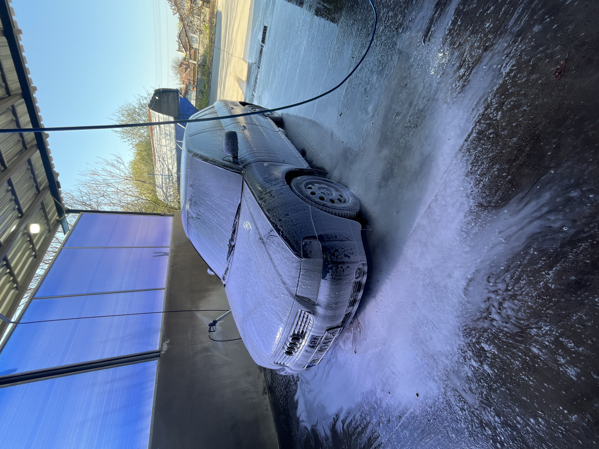 A dark car is covered in white foam at a car wash in Glojene, Bulgaria. The car is parked inside a structure with a corrugated metal roof and translucent blue panels along the sides. A black hose drapes across the top of the car. Foam and water are accumulating on the ground. The car's rear door is open. The car’s license plate reads "BH 1494 CF”. Bare trees are visible in the background.