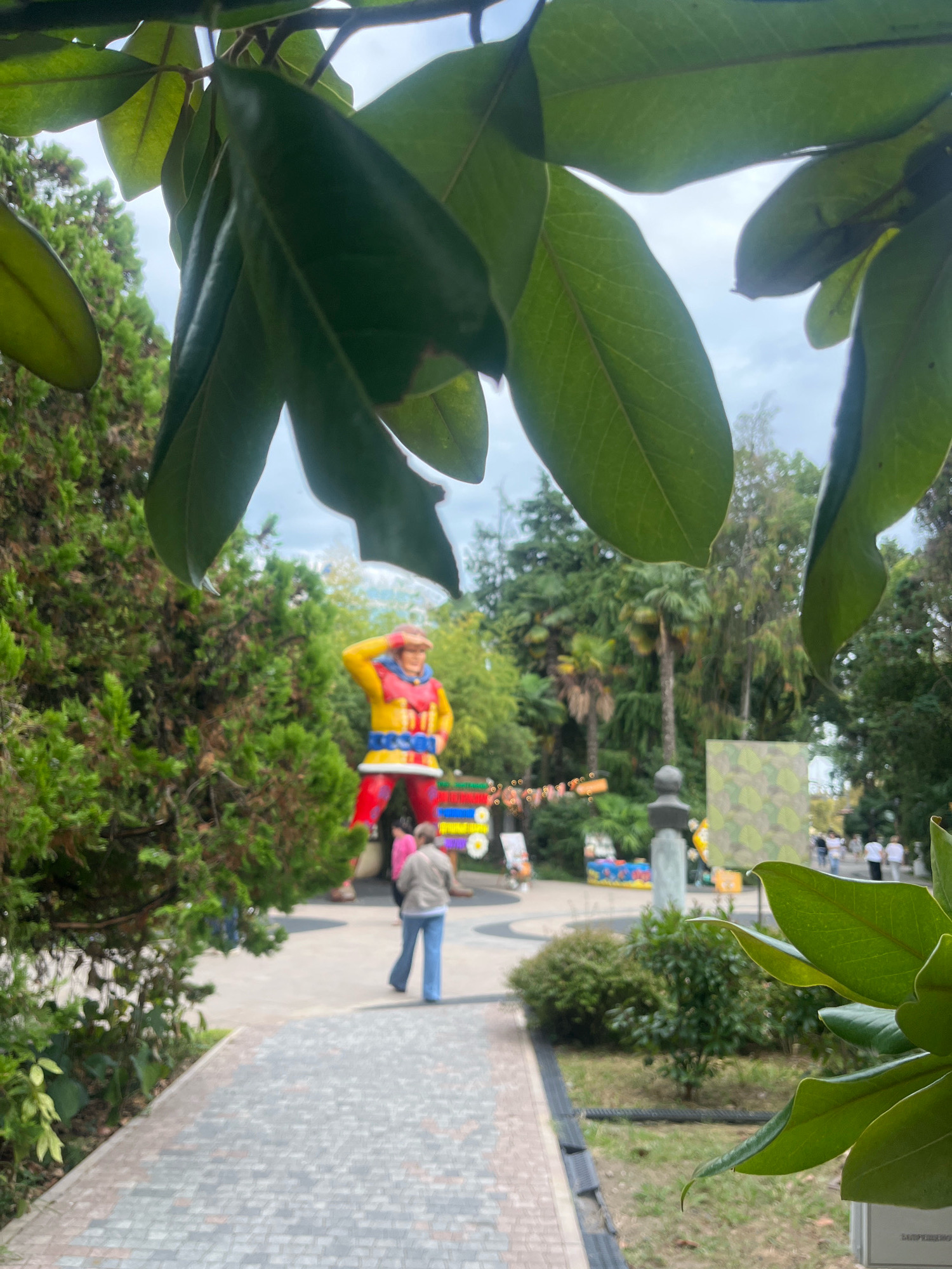 The image depicts an outdoor scene, likely in a park or public space. In the foreground, large green leaves obscure part of the view. In the mid-ground, a person in a light brown jacket and blue pants walks away from the camera along a paved pathway. Behind this person, a large, colorful statue of a figure in a red, yellow, and blue costume stands prominently. The statue appears to be a playful character, possibly a jester or a clown, with a red hat. In the background, lush greenery, including palm trees and other foliage, suggests a warm climate. The sky is overcast, with a light gray cloud cover. The pathway is made of gray and reddish-brown pavers arranged in a pattern. Further in the background, other people are visible walking, and there are some decorative elements and signs, including a camouflage-patterned board. The presence of palm trees and the style of the park suggest a location in Sochi, Russia, potentially a resort area or an amusement park.