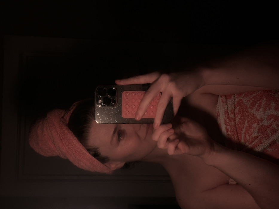 The image captures a mirror selfie taken indoors in low light. The primary subject is a person, likely female, whose reflection is seen in the mirror. The person is holding a smartphone with both hands, positioned to capture their reflection.
The person is wearing a soft, coral or pink-colored towel wrapped around their head, covering their hair. Another towel, with a white background and a coral/orange patterned design (possibly floral or geometric), is draped around their torso, visible on the right side of the frame. Their skin appears fair. Due to the phone's position, only one eye and parts of their face are clearly visible, obscured slightly by the phone and hands. The visible eye is light-colored.
The smartphone has a silver or clear case that appears to have glitter or speckles embedded in it. It features a prominent camera bump with multiple lenses on the back. Attached to the back of the phone case is a coral or pink-colored accessory with an array of circular indentations, possibly a PopSocket or a fidget toy.
The lighting in the scene is dim, casting a warm, slightly reddish hue on the person and the towels. The background reflected in the mirror behind the person is very dark, suggesting minimal illumination in the room beyond the subject themselves, or a dark wall. No specific details of the room are clearly discernible due to the darkness, except for what appears to be a faint outline of a door frame or wall molding in the very dark upper left corner of the original image. No text is visible in the image.