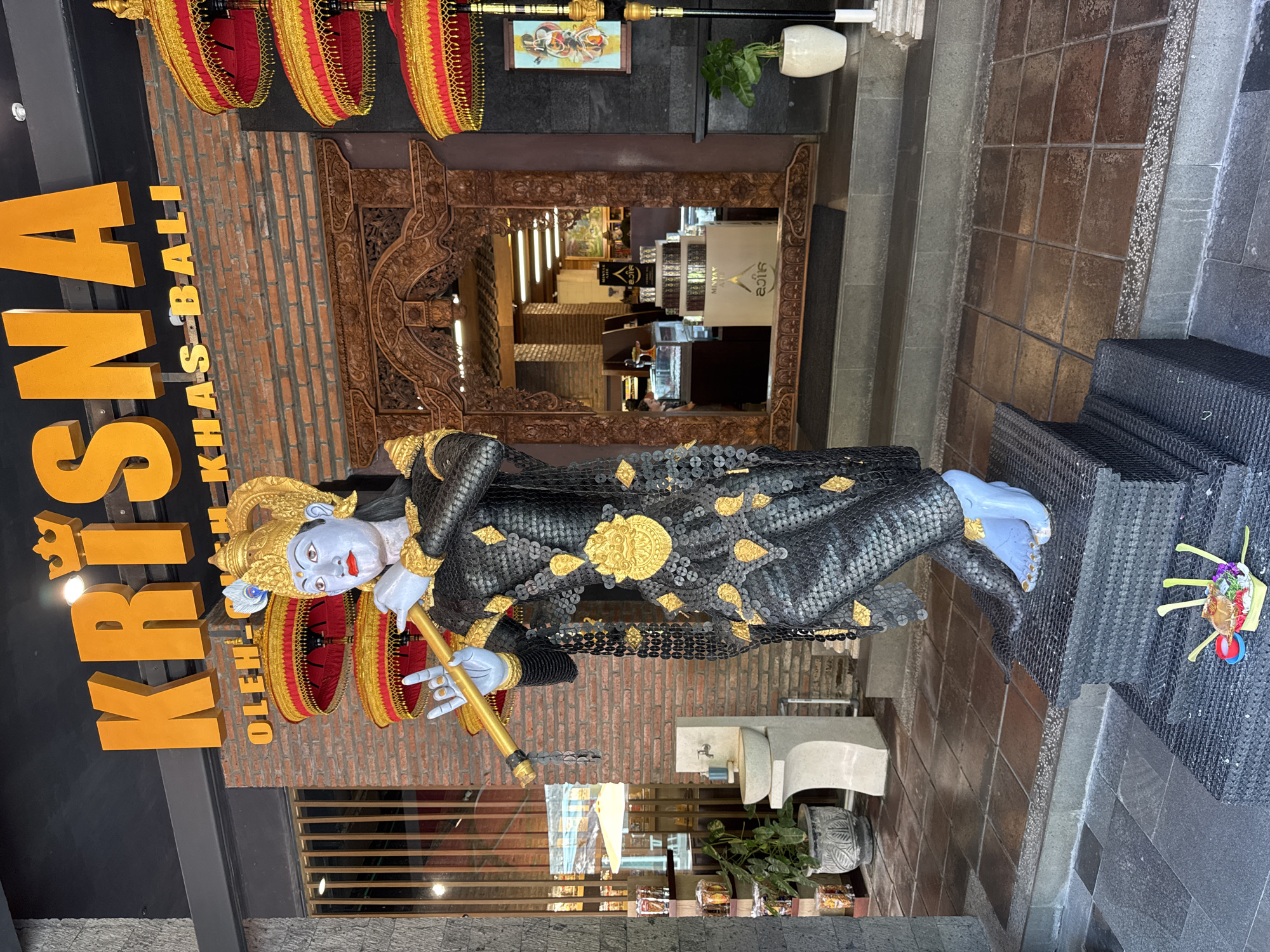 The image is taken outdoors, likely in the city of Kuta Selatan, Indonesia, depicting the entrance of a commercial establishment. A large, colorful statue of Krishna is positioned prominently in front of the building. Krishna is depicted with a blue complexion, elaborate headdress, and ornate clothing, playing a flute. He is adorned with gold embellishments.
To the left of the statue, a sign reads "KRISNA OLEH OLEH KHAS BALI" in large, yellow block letters. The building's facade is decorated with traditional Balinese elements, including a carved wooden frame around the doorway, and red and gold decorative pieces hanging above.
A set of steps leads up to the entrance. A planter with greenery is visible beside the steps. The area appears to be well-maintained and features brick and tile flooring. The doorway provides a glimpse into the interior of the establishment, where shelves, lighting fixtures, and staff can be seen. A water feature appears between the statue and the door way.