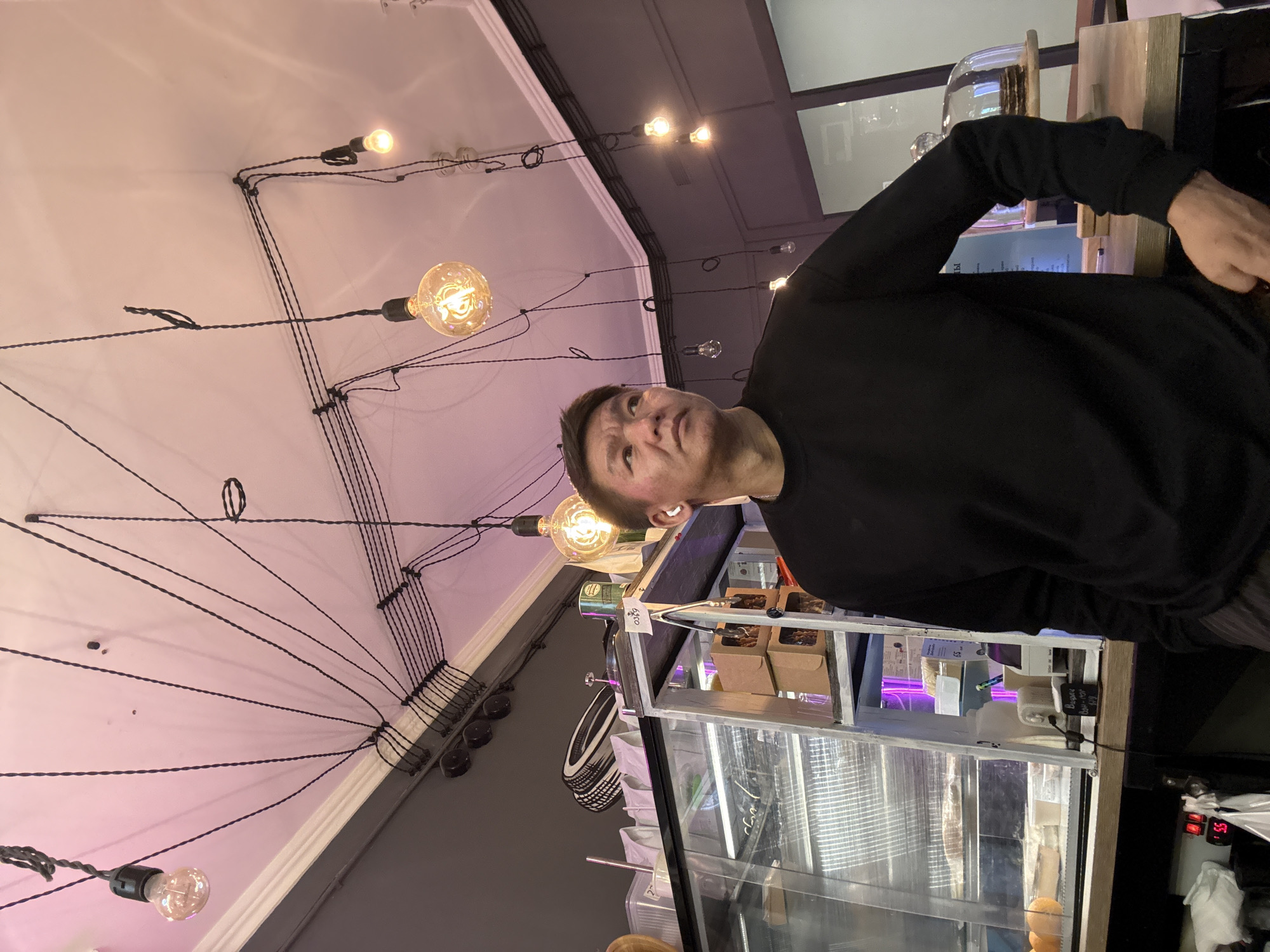 A man with short dark hair and light brown skin stands in what appears to be a cafe. He is wearing a black crew neck long-sleeved shirt and dark pants. He has a white earbud in his left ear and is looking to his left. Behind him, a refrigerated display case is filled with food items and shelves above it hold bags and containers. Above the man, exposed light bulbs hang from black cords connected to the ceiling. The ceiling itself is painted pink and grey, with a white decorative molding. The scene is illuminated by the warm glow of the light bulbs and ambient lighting from the display case. The cafe environment is suggested by the display of food and the casual atmosphere. The location is not explicitly stated to be Moscow, Russia, but the presence of Cyrillic text on signage within the image indicates a Russian-speaking environment.