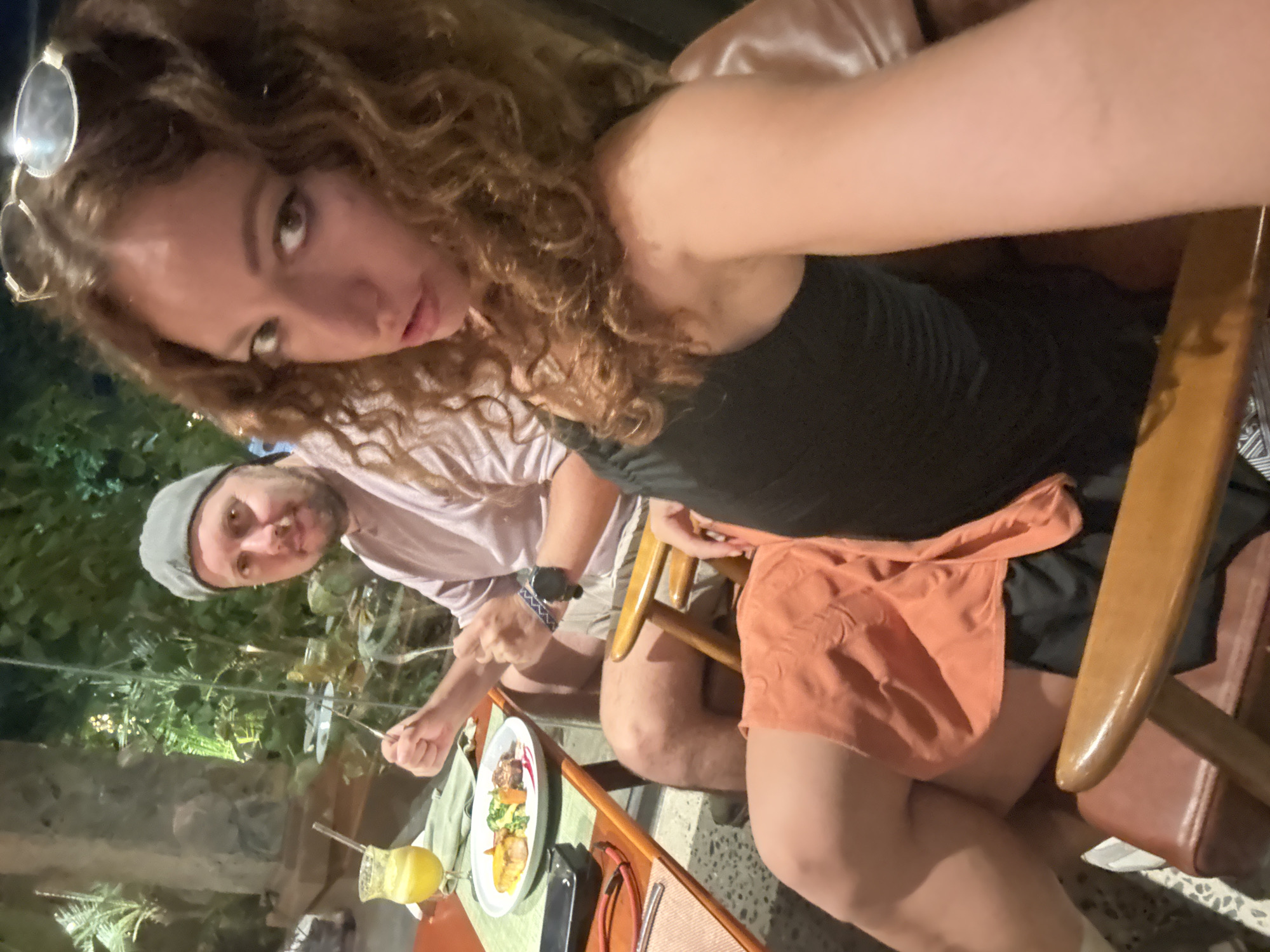 In a dimly lit restaurant, a man wearing a gray cap and a pink shirt sits at a table, holding a fork and knife. In front of him is a plate of food with salmon, vegetables, and sauce, alongside a glass of orange juice. To his right, a woman with curly brown hair and sunglasses on her head poses for the camera. She wears a black top and an orange skirt. The background shows lush greenery illuminated by soft lights, suggesting an outdoor or garden setting. The image is taken at a close-up angle, focusing on the two individuals and their immediate surroundings.