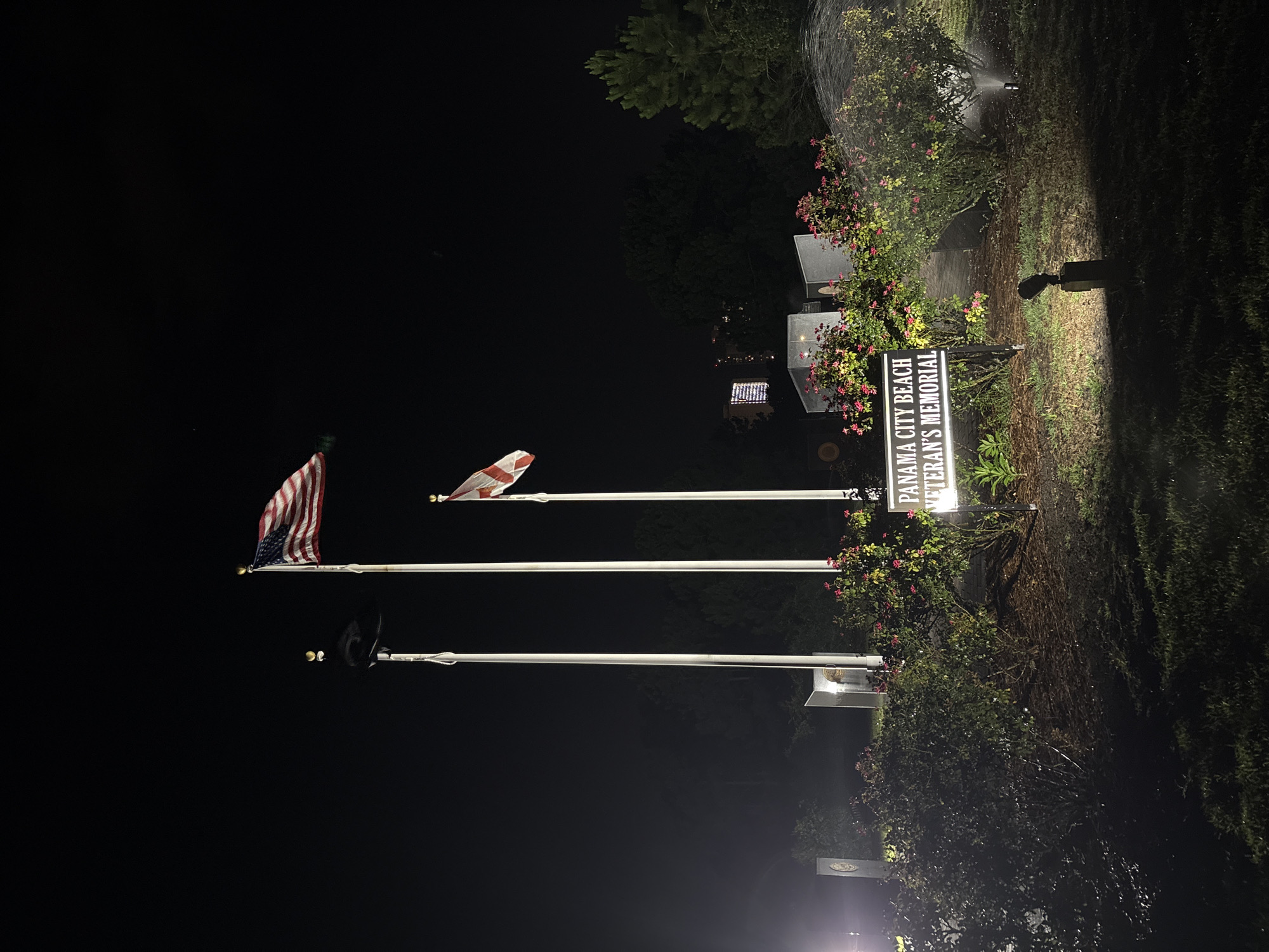 This media file depicts an outdoor scene at night, illuminated by artificial lights and a sprinkler system that appears to be in operation, casting a misty spray. The primary subjects are three flagpoles, two of which are flying flags. The topmost flag is the American flag, with its stars and stripes clearly visible. Below it, another flag is partially visible, appearing to be white with red crosses. The third flagpole is empty.
The setting is a memorial area, as indicated by a lit sign that reads "PANAMA CITY BEACH VETERAN'S MEMORIAL". Behind the sign and flags, there are structures that resemble memorial plaques or monuments, some of which are also illuminated. Lush green bushes and trees surround the area, with some flowering plants adding touches of pink to the foliage. The ground appears to be grass and dirt, wet from the sprinkler.
The time of day is clearly night, with the darkness contrasting with the illuminated elements. The atmosphere is somber and respectful, befitting a memorial site. The presence of the sprinkler suggests that the area is being maintained, possibly to water the plants or to clean the surfaces. The overall scene conveys a sense of remembrance and honor for veterans.