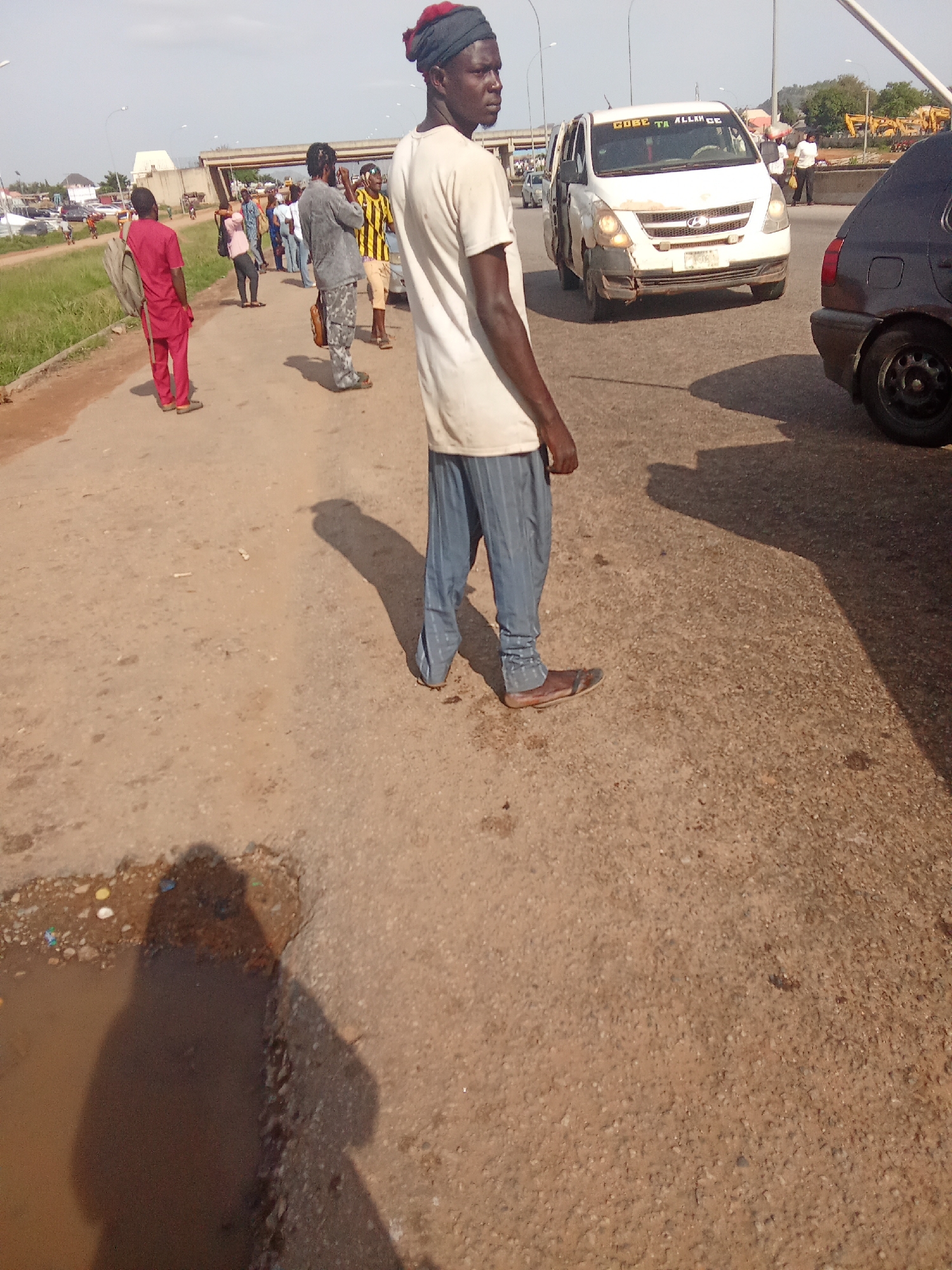 A man wearing a red garment stands on a paved road in an outdoor urban environment. The road is a mix of asphalt and gravel, with a puddle of water on the left side. In the background, other people are visible, as are several vehicles, including a white van with text on the windshield and a dark car. An overpass or bridge is also present in the distance, along with construction equipment. The sky is clear and bright.