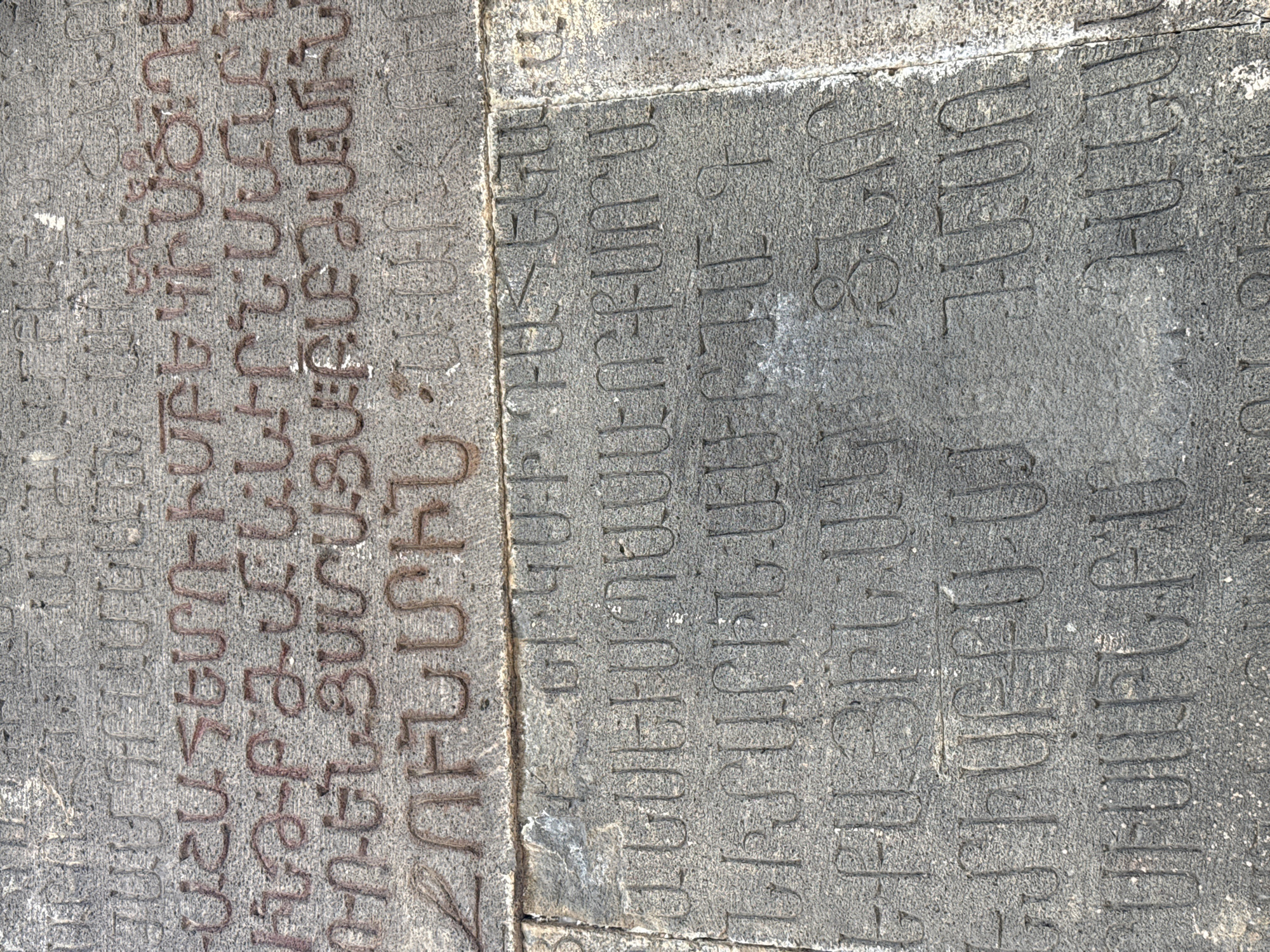 The image shows an inscription in Armenian script carved into a stone surface. The script consists of a series of vertical lines with rounded or angular elements attached, appearing as raised carvings. The stone is weathered and textured, with some discoloration, possibly due to age and exposure. The inscription is divided into several lines of text. This stone is located in the village of Goght, Armenia, and is likely a historical or religious monument.