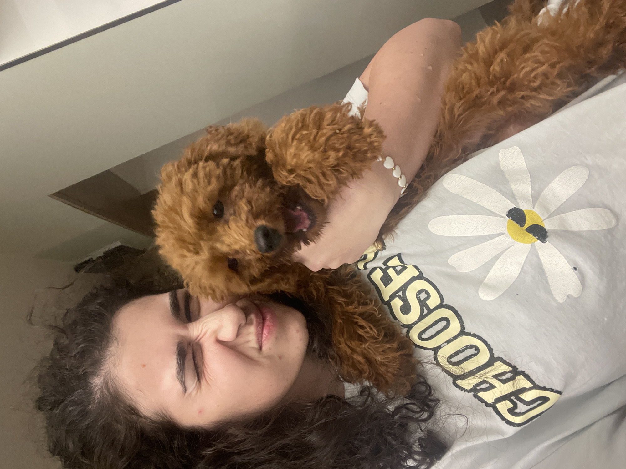 A close-up, playful portrait shows a young woman and a small, reddish-brown poodle engaging in an affectionate interaction, likely indoors in Sofia, Bulgaria. The image is oriented with the woman's head to the bottom left and the dog's head near the center.
The woman, with dark curly hair, is lying down and looking at the camera with a playful, scrunched-up expression, her eyes slightly closed and lips pursed. Her right arm is visible, adorned with a white bracelet featuring heart-shaped beads. She is wearing a light-colored t-shirt with a prominent white daisy design with a yellow and black center. Below the daisy, the word "CHOOSE" is clearly visible in bold yellow letters with a dark outline.
The fluffy, reddish-brown poodle is positioned close to the woman's face, with its mouth slightly open, suggesting a moment of playfulness or a vocalization like a bark or whine. The dog has dark eyes and a dark nose. Its fur appears thick and curly. The woman's arm is gently wrapped around the dog, indicating a close bond.
The background is simple and uncluttered, appearing to be a plain light-colored wall or ceiling, suggesting an indoor setting. The lighting is soft, highlighting the intimate and joyful nature of the scene. The overall mood of the image is one of happiness, affection, and lightheartedness between the woman and her canine companion.