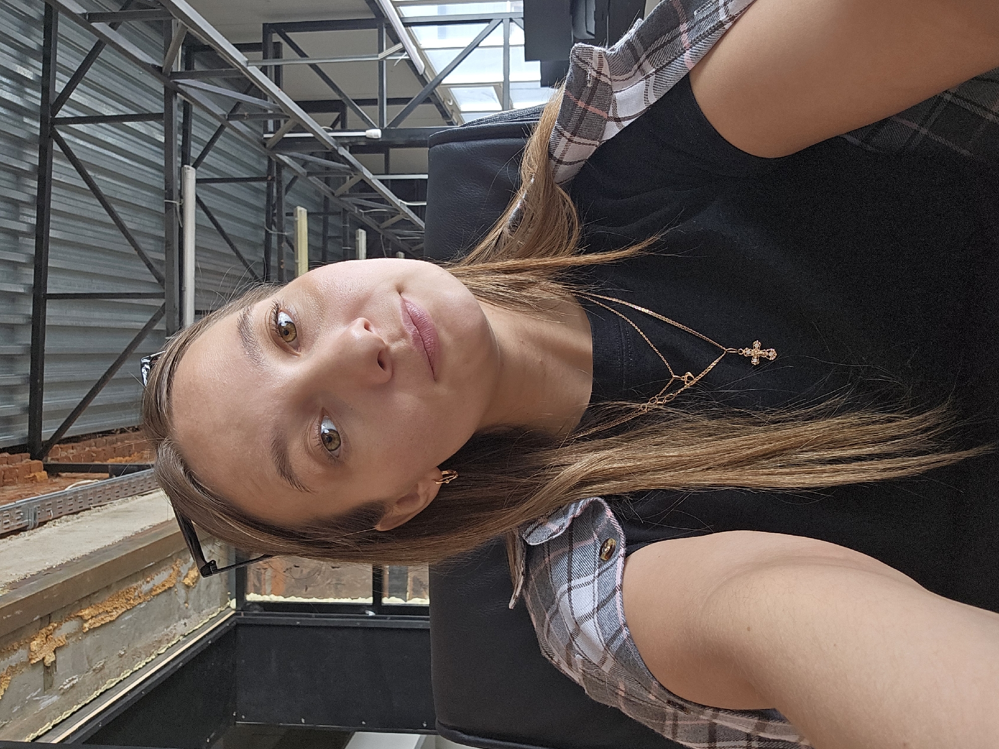 The image captures a young adult female in an indoor setting in the city of Chaikovsky, Russia. The subject has long, light brown hair and light-colored eyes, looking directly at the camera with a neutral expression. She is wearing a black t-shirt, with a grey, white, and pink plaid flannel shirt draped over her shoulders. Visible accessories include small gold hoop earrings and two gold necklaces, one with a heart pendant and another featuring a stone-set cross pendant. She is seated in a black chair, and her right upper arm is visible on the lower right, suggesting she is holding the camera for a selfie. The background reveals an industrial-style interior, characterized by an exposed metal truss ceiling with corrugated panels, and fluorescent light fixtures. Elements of exposed brickwork with foam insulation and dark metal structural framing are also visible. Windows are present on the right side, providing ambient light.