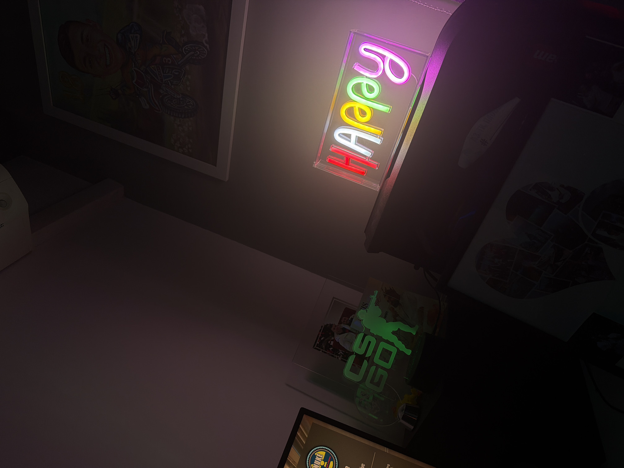The image shows a dimly lit room with various objects visible. On the left wall is a framed picture of a person on a motorcycle. Next to it is a neon sign that says "Happy" in rainbow colors. On the right, there is a dark object, possibly a printer, next to a framed picture with a heart-shaped collage. Below, there is another neon sign, green in color, and what appears to be a monitor displaying a logo. The location appears to be indoors, possibly a bedroom or office. The city and country are not identifiable from the image.