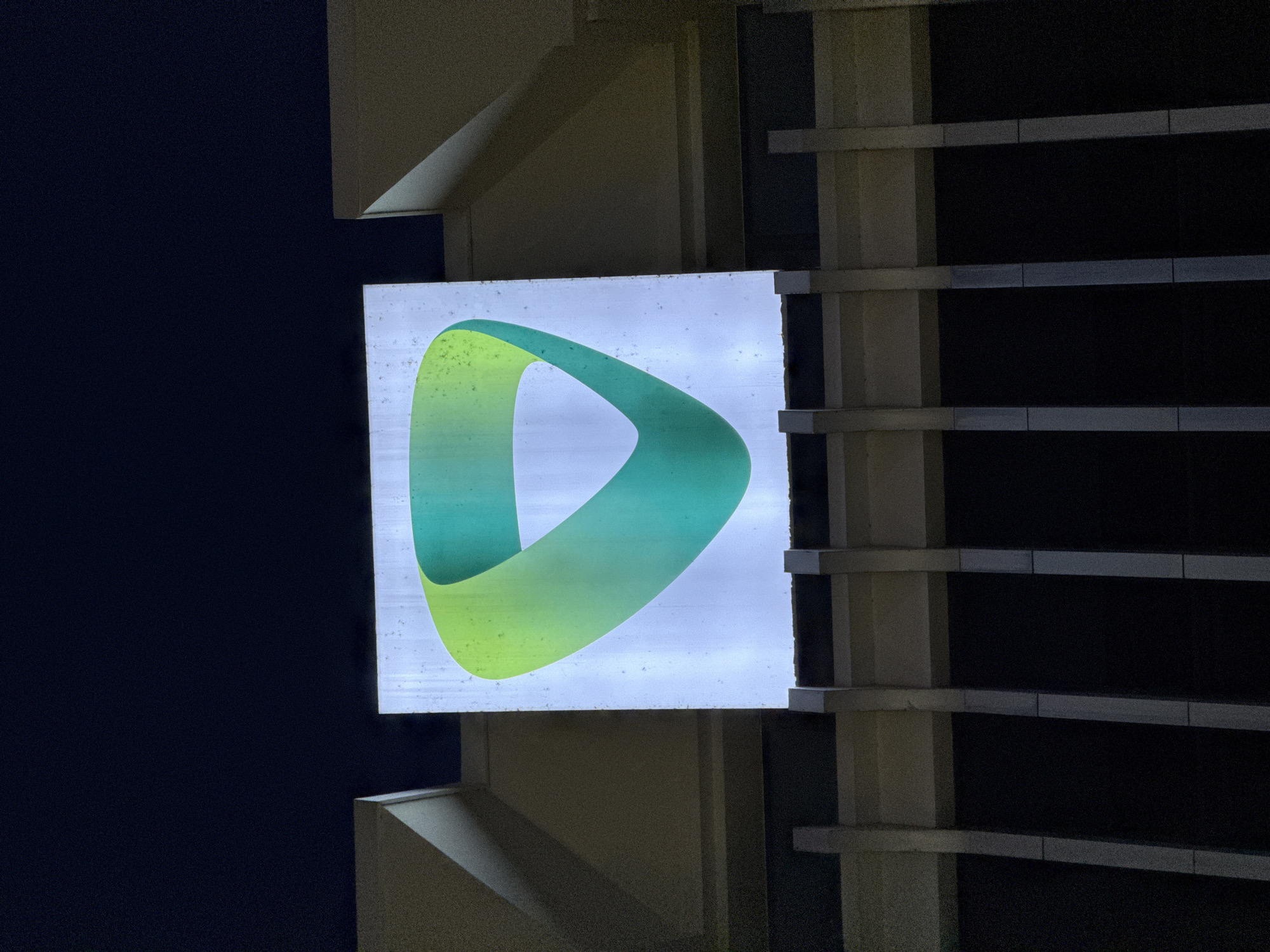 The image shows a close-up view of a lit-up square sign on the facade of a building at night. The sign features a stylized, curved green and yellow logo that appears to be a Möbius strip or a similar continuous loop. The building's architecture is visible, with geometric shapes and vertical elements suggesting a modern design. The background is a dark, clear night sky. The image does not contain any people or specific identifiable landmarks of Nha Trang, Vietnam.