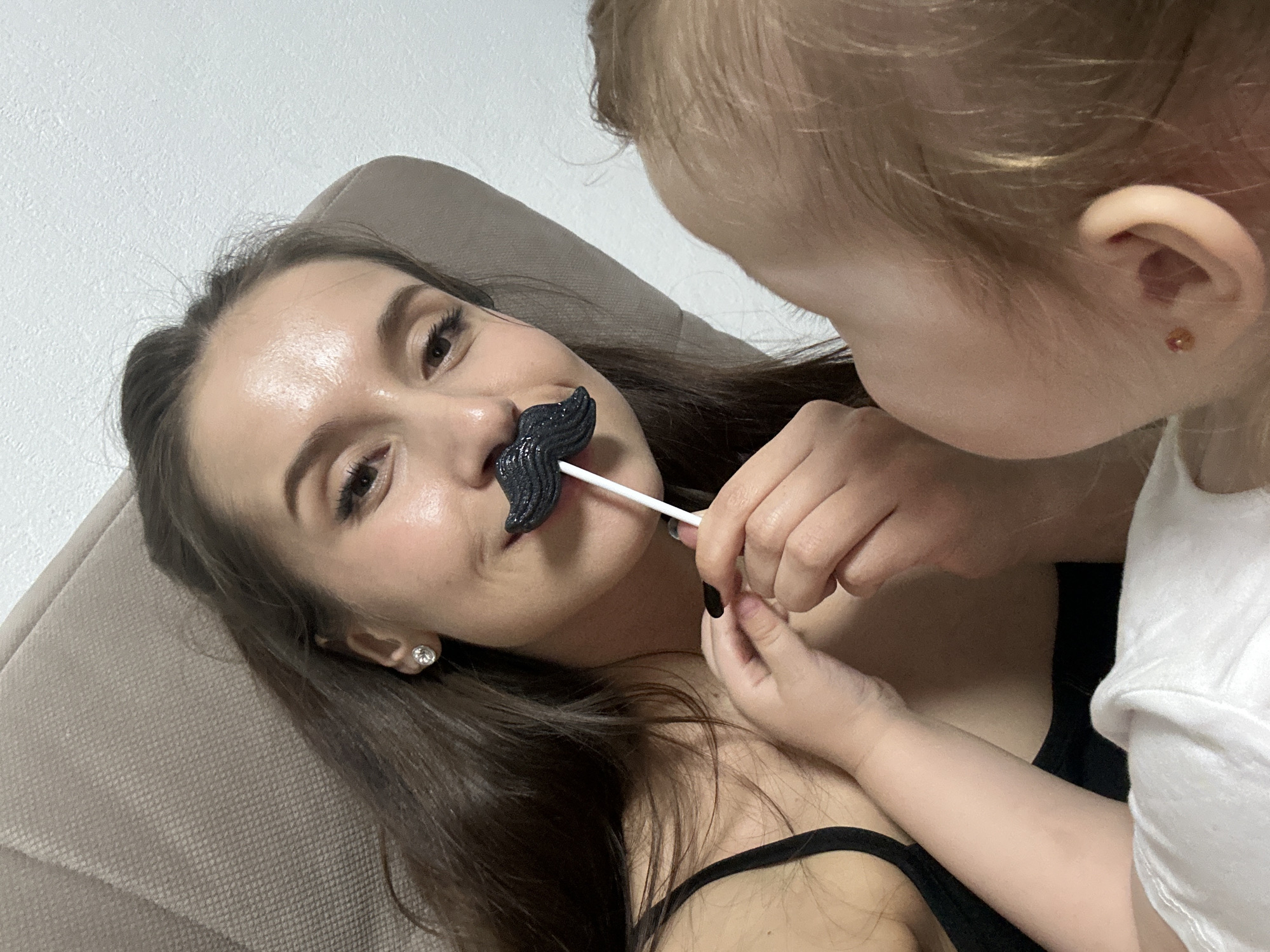 A woman is lying down on a couch or cushion, and a young child is holding a candy mustache on a stick up to her face. The woman is fair-skinned with brown hair and is wearing a black tank top and small diamond-shaped earrings. She is looking towards the child with a smile. The candy mustache is black and textured. The child is fair-skinned with light hair and is wearing a white t-shirt. The child's right ear is visible, with a small red earring. The background is a plain white wall. The specific location is not discernable from the image and cannot be confirmed as Yekaterinburg, Russia.