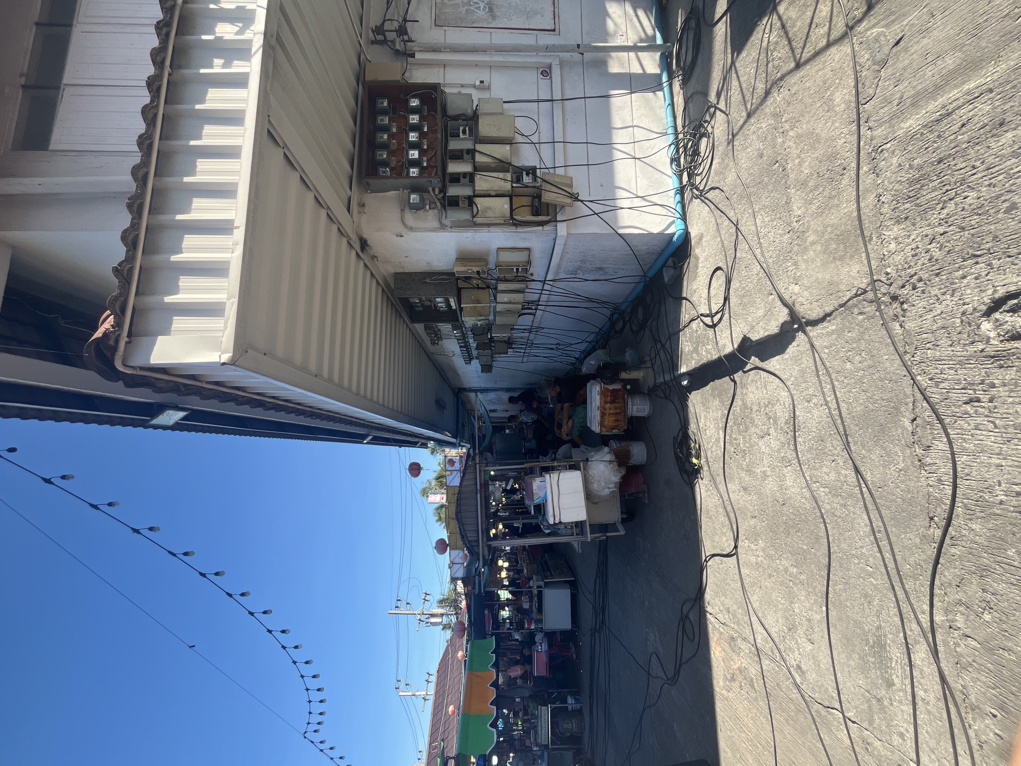 The image depicts a street market scene in Tambon Ko Pha Ngan, Thailand, under a clear blue sky. In the foreground, the ground is paved with concrete, crisscrossed by numerous black electrical cables, some coiled and others spread out. To the right, a white building is visible, featuring a series of outdoor electrical meter boxes mounted on the wall, with many cables connected to them. The building has a white corrugated metal roof overhang.
In the background, the market stalls are set up along the street. Some stalls have colorful awnings in green, orange, and yellow. Red lanterns are hanging from some structures, and strings of decorative lights are stretched across the sky. People can be seen at the market, with some individuals sitting and others appearing to be tending to their stalls or browsing. The overall atmosphere suggests a bustling daytime market.